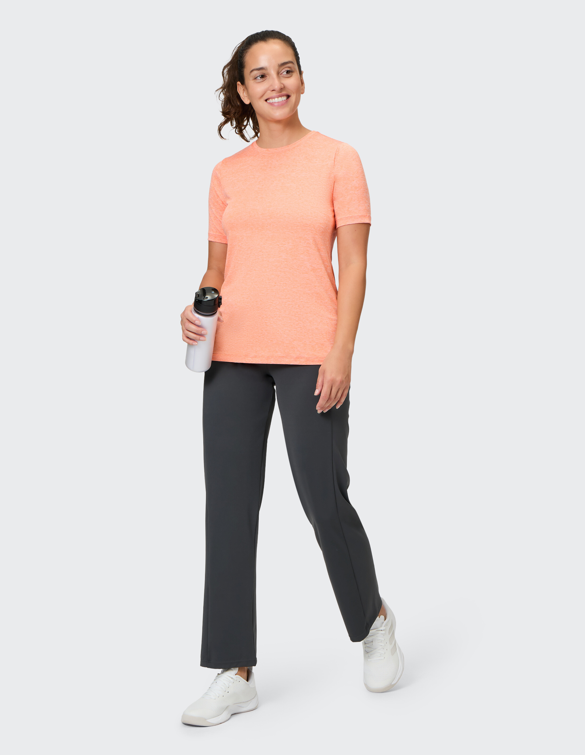 Frau in einem korallenroten T-Shirt und einer grauen Hose, hält eine Wasserflasche in der Hand und lächelt.