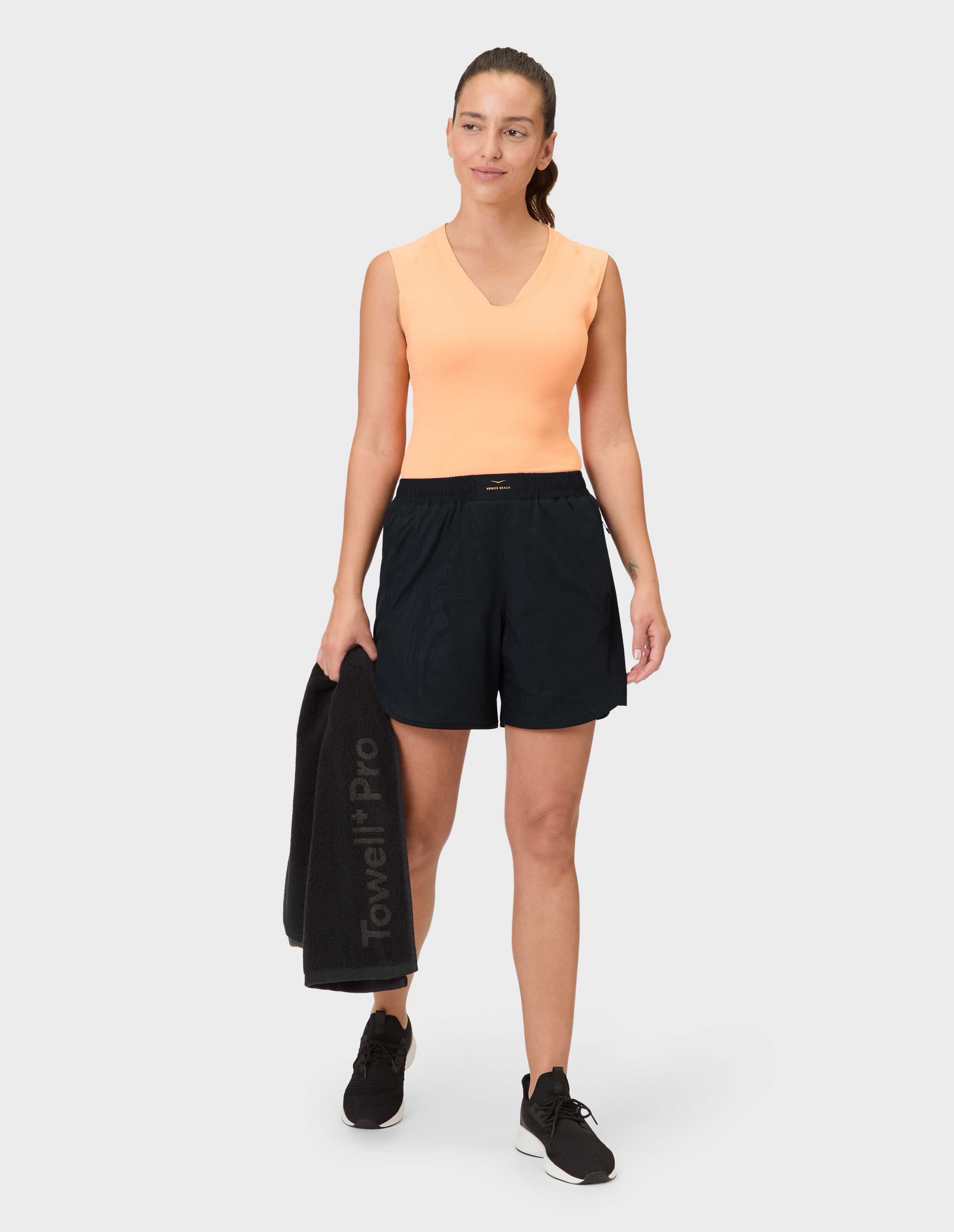 Frau in orangefarbenem ärmellosem Top und schwarzen Shorts, mit einem Handtuch in der Hand, bereit für Freizeitkleidung.