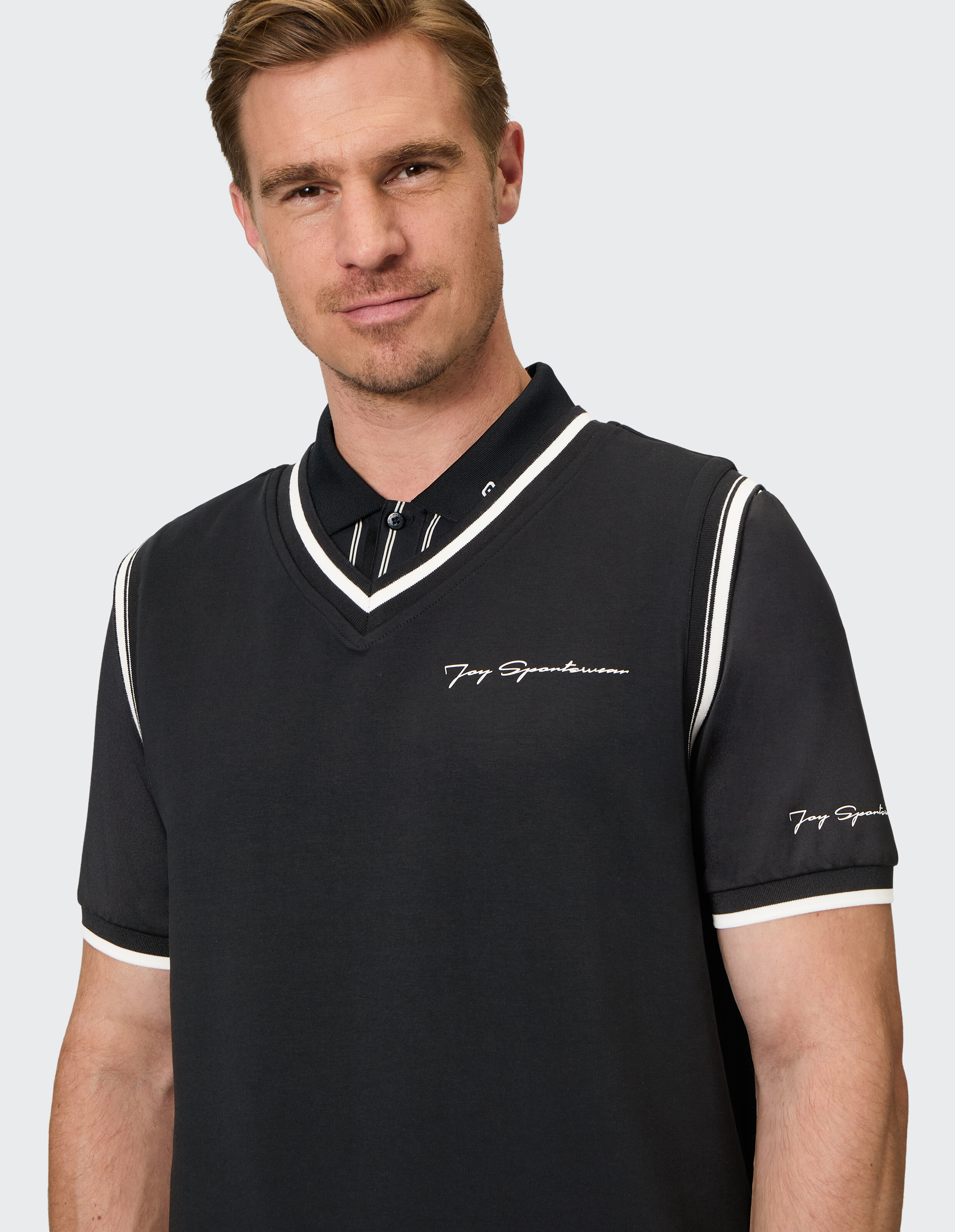 Mann mit kurzem Haar trägt ein dunkelblaues Polohemd mit weißem Saum und Aufschrift Joy Sportswear, steht vor grauem Hintergrund.