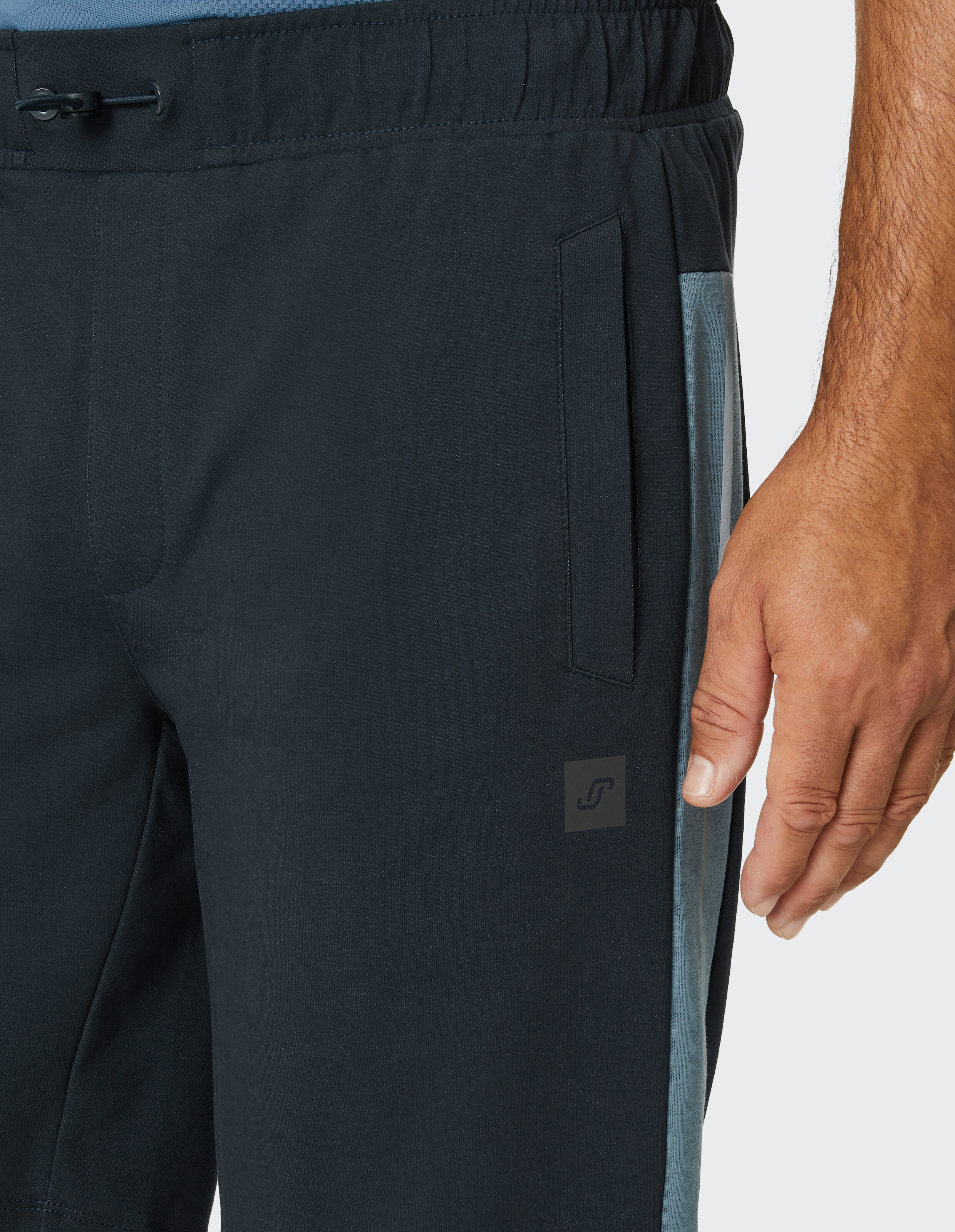 Nahaufnahme einer schwarzen Jogginghose mit grauen Seitenstreifen, Kordelzug und einem Logo, getragen von einer Person mit nacktem Arm.