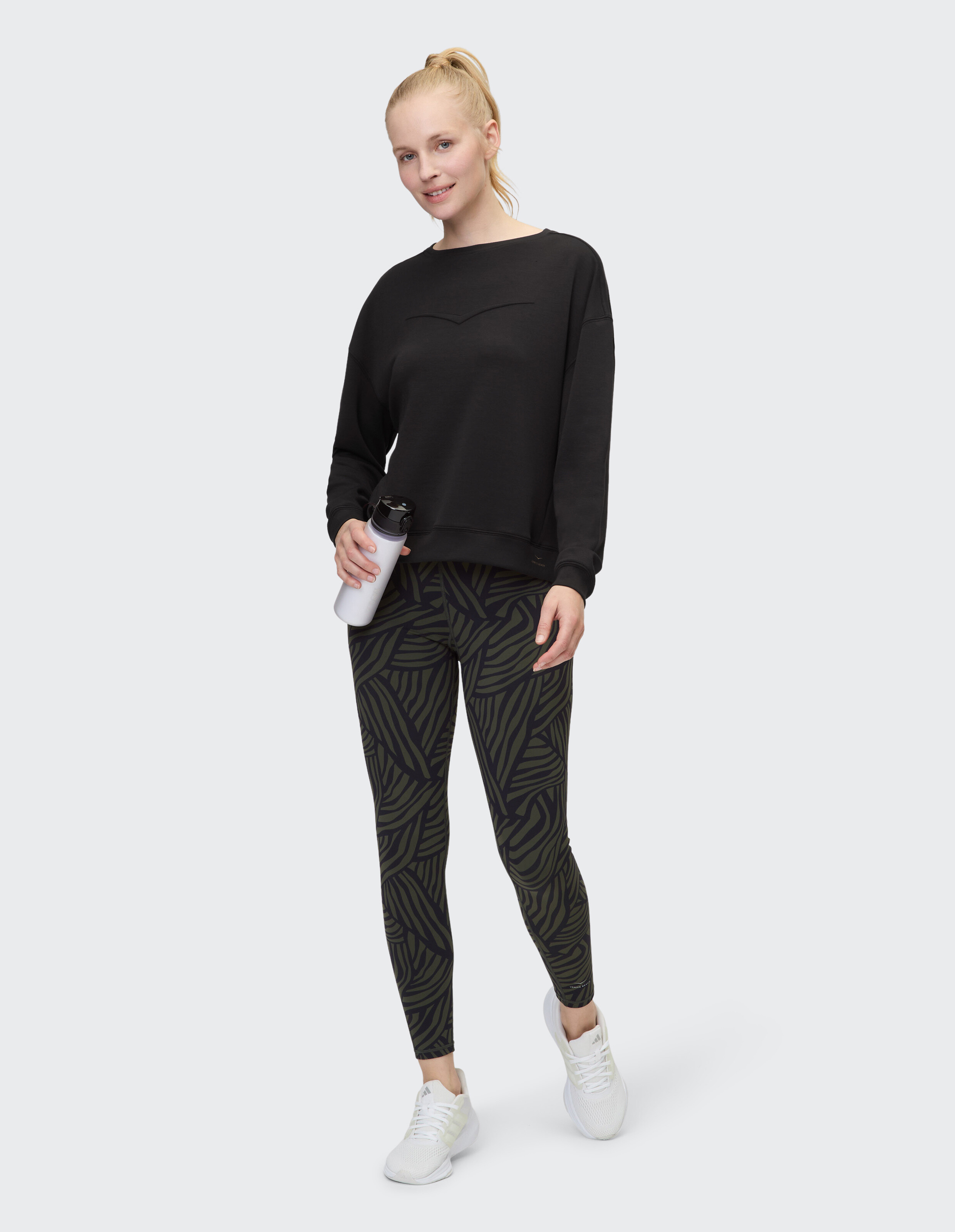 Frau in schwarzem Sweatshirt und gemusterten Leggings, hält eine Wasserflasche in der Hand.
