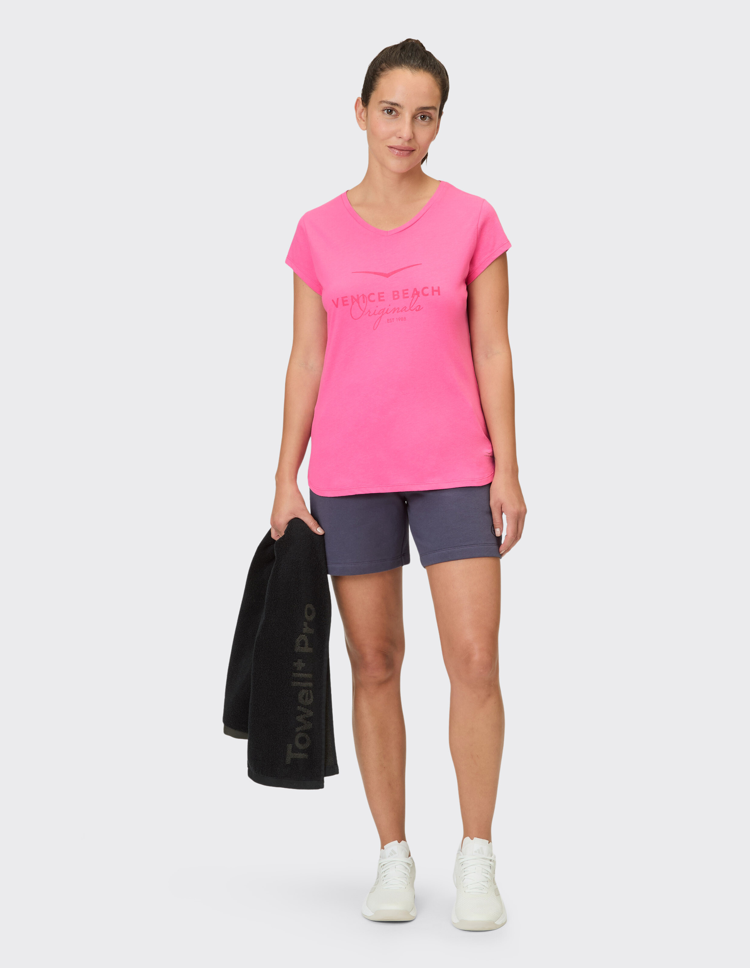 Frau in einem rosafarbenen Venice Beach-T-Shirt und dunkelblauen Shorts, mit einem schwarzen Handtuch in der Hand.