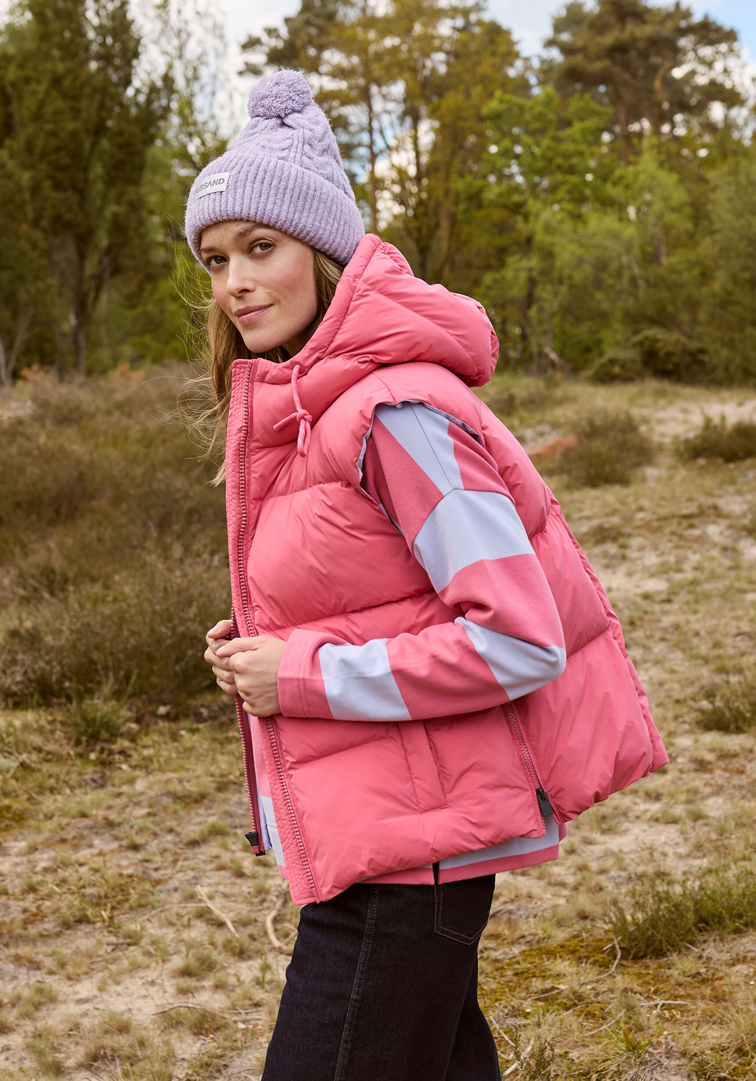Frau in rosa Pufferweste und gestreiftem Oberteil, trägt eine lavendelfarbene Mütze im Freien.