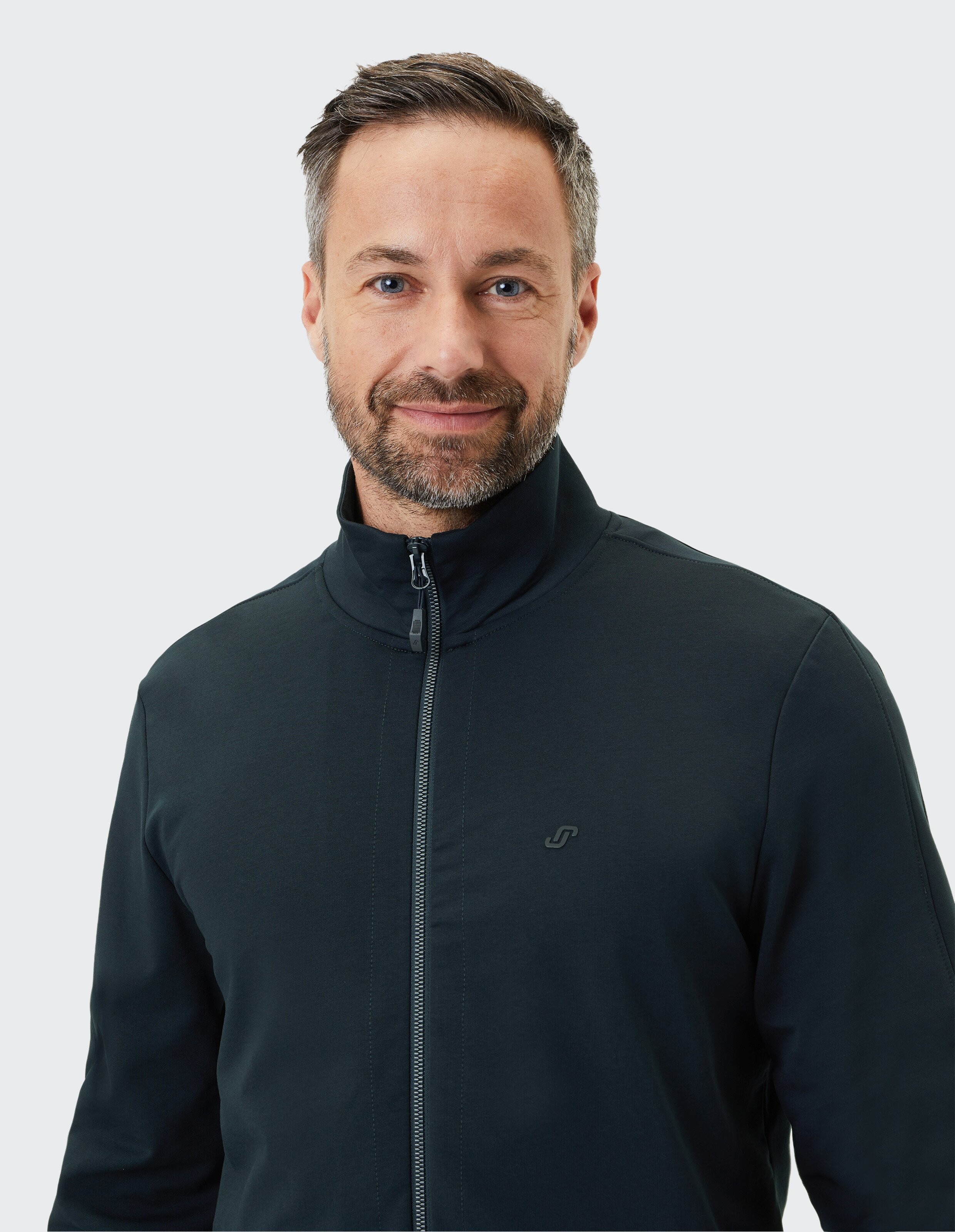 Ein lächelnder Mann mit Bart trägt eine dunkelblaue Jacke mit Reißverschluss und steht vor einem hellgrauen Hintergrund.