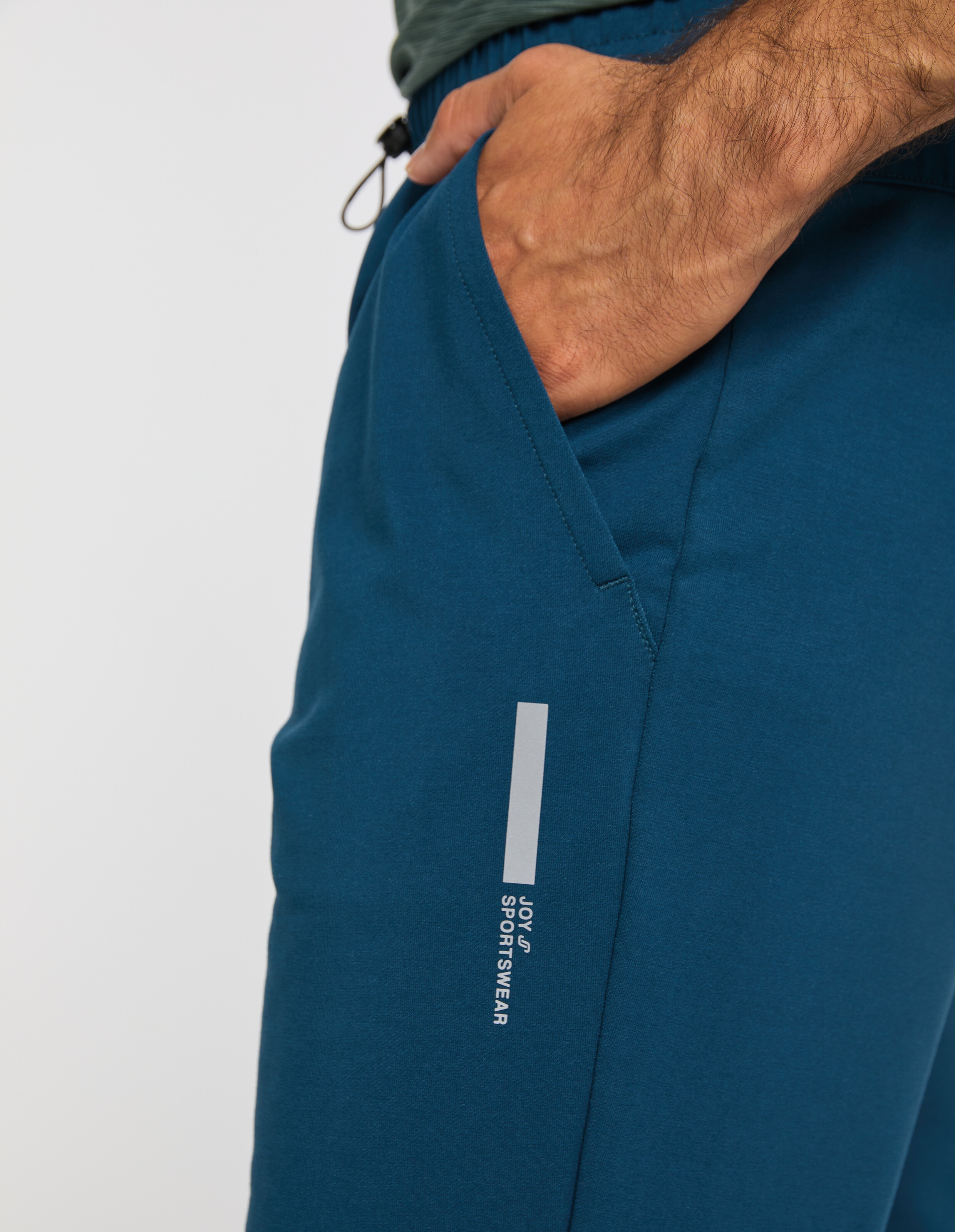 Ein Mann trägt eine blaue Sporthose mit seitlicher Tasche und reflektierendem Aufdruck JOY SPORTSWEAR, Hand in der Tasche.