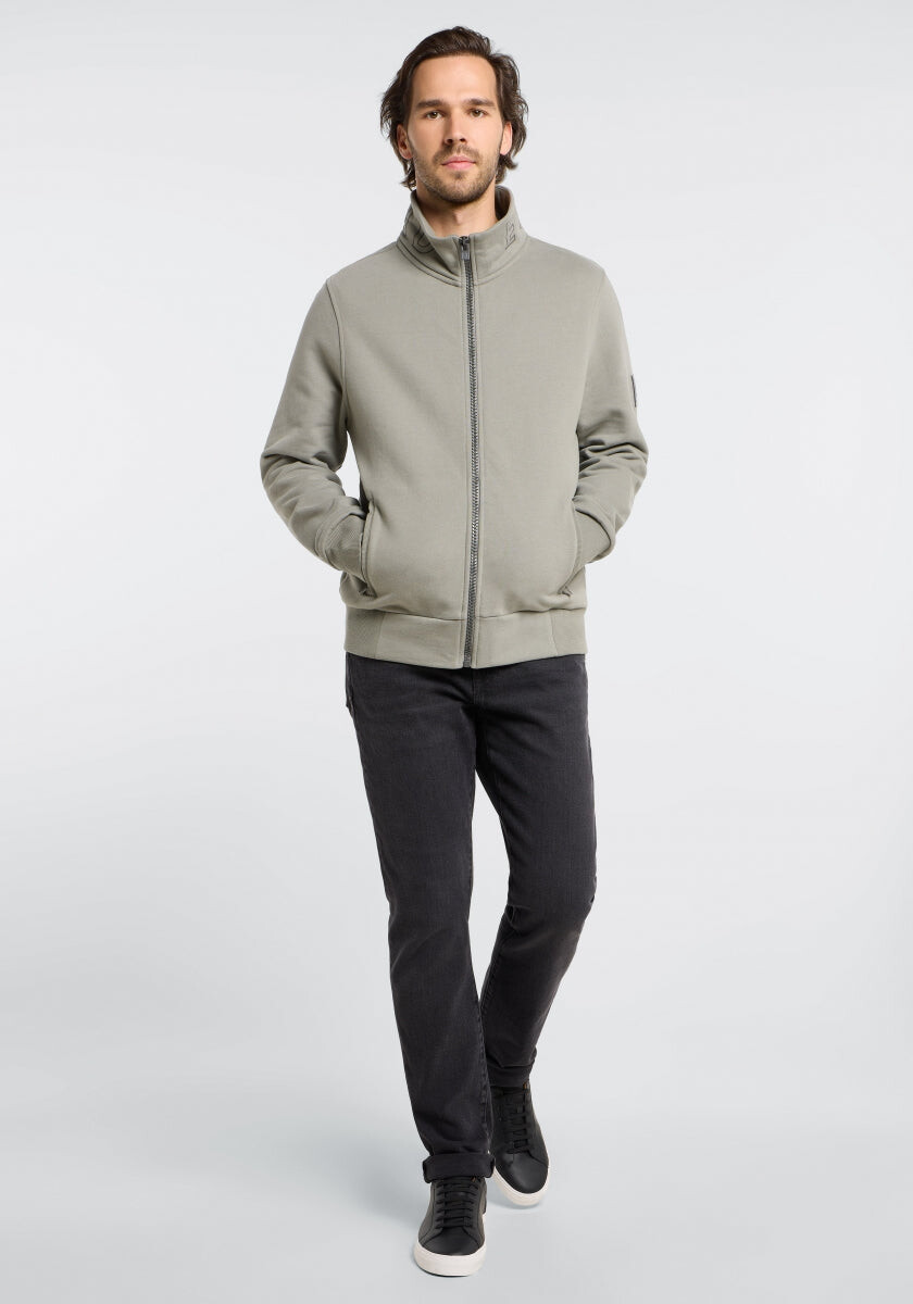 Mann in einer hellgrauen Jacke mit Reißverschluss und dunklen Jeans, gestylt für Freizeitkleidung.