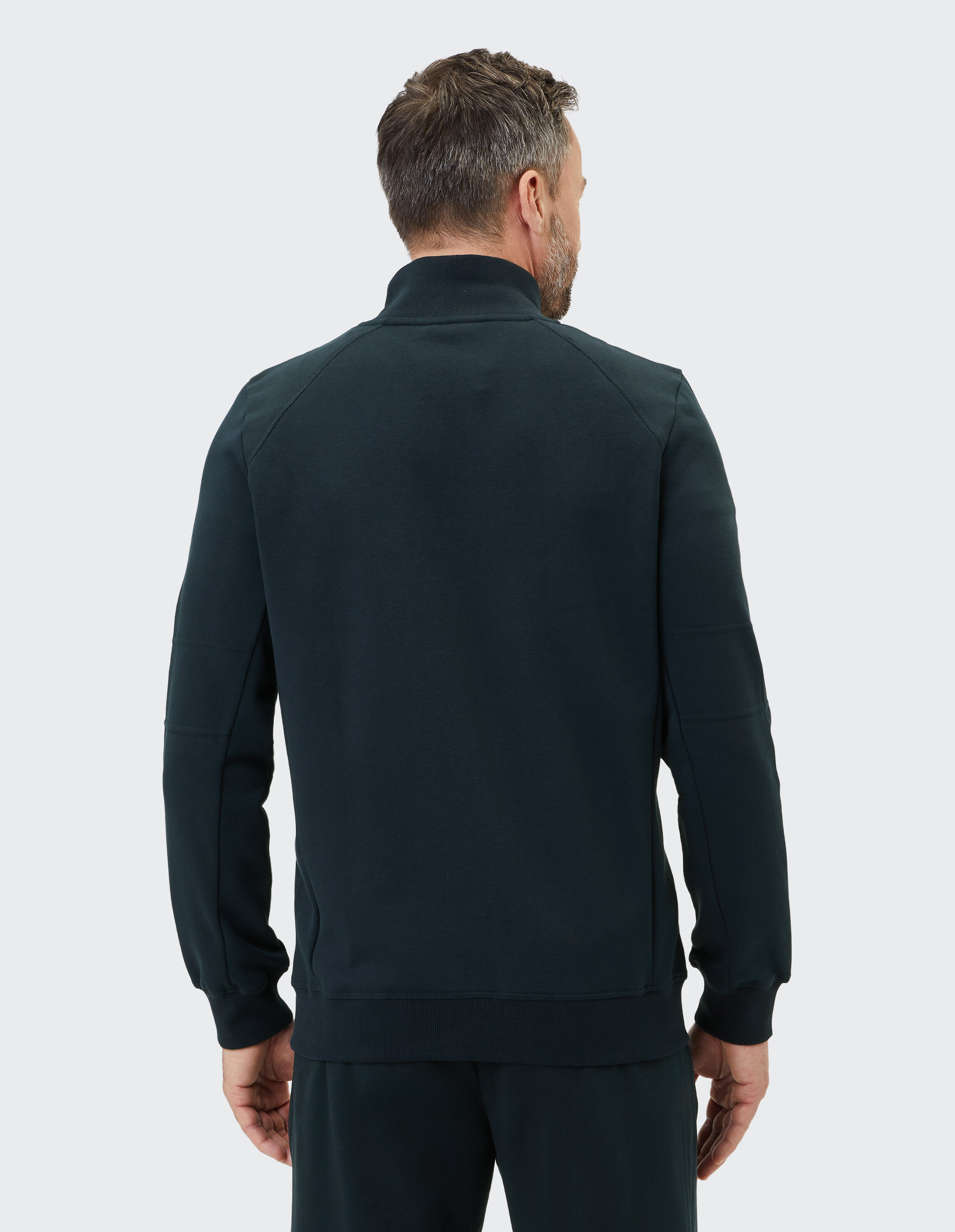 Rückansicht eines Mannes in dunkelblauer Sweatshirtjacke mit hochgeschlossenem Kragen, er steht vor einem hellen Hintergrund.