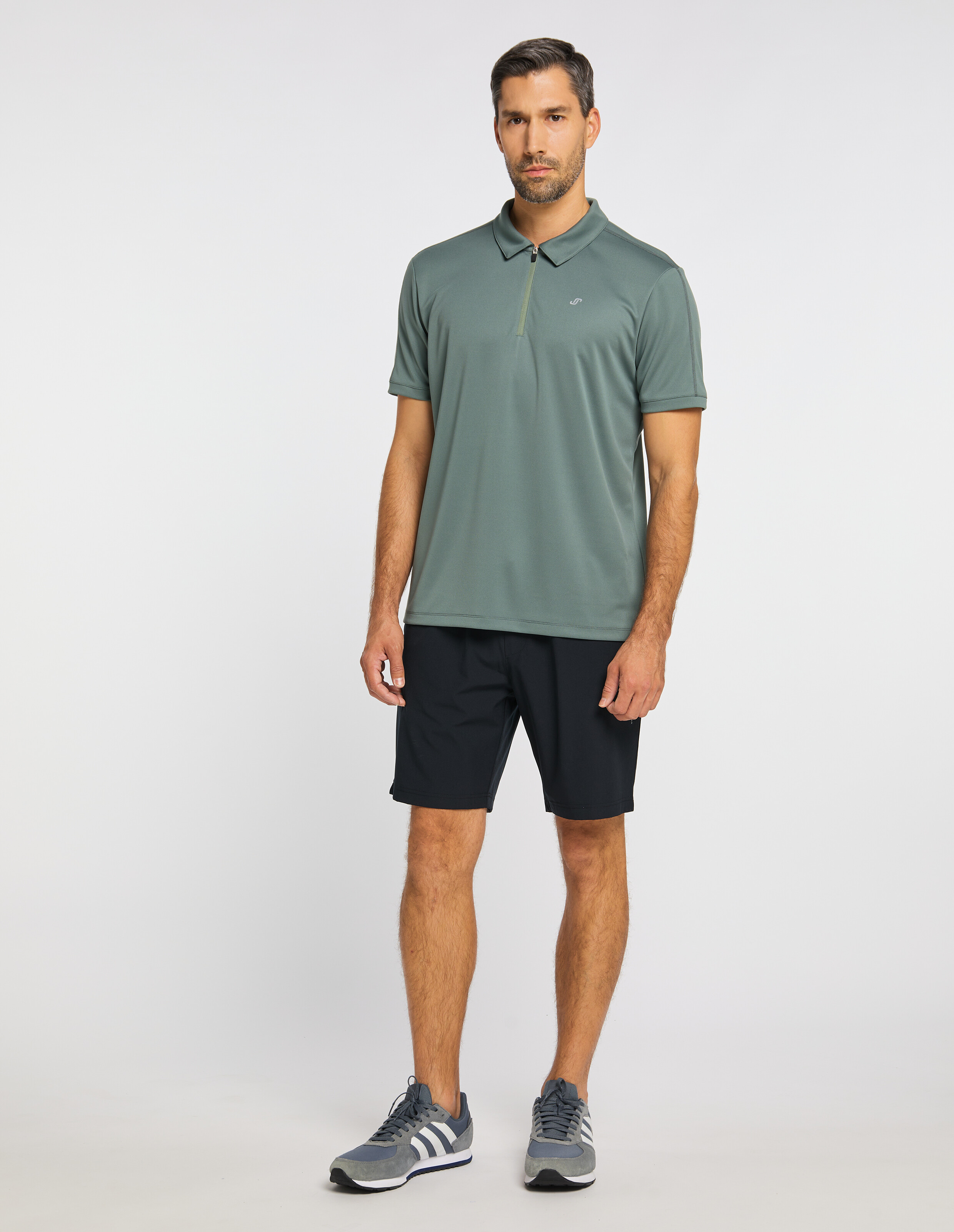 Ein Mann in grünem Poloshirt und dunklen Shorts steht vor neutralem Hintergrund