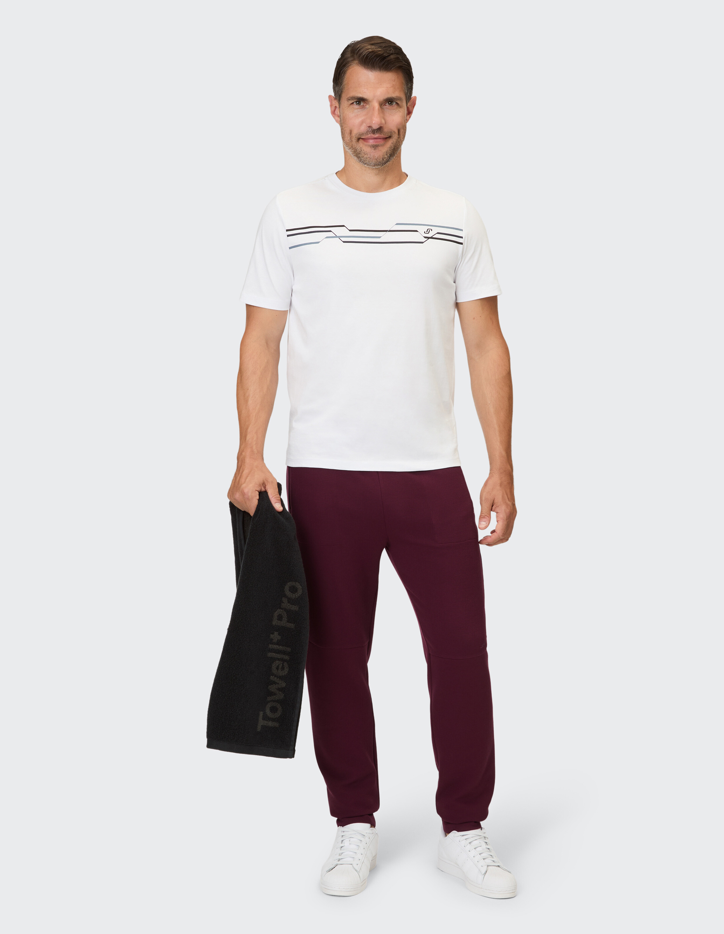 Mann in lässigem weißem T-Shirt mit Streifen und burgunderfarbener Jogginghose, hält ein Handtuch in der Hand.
