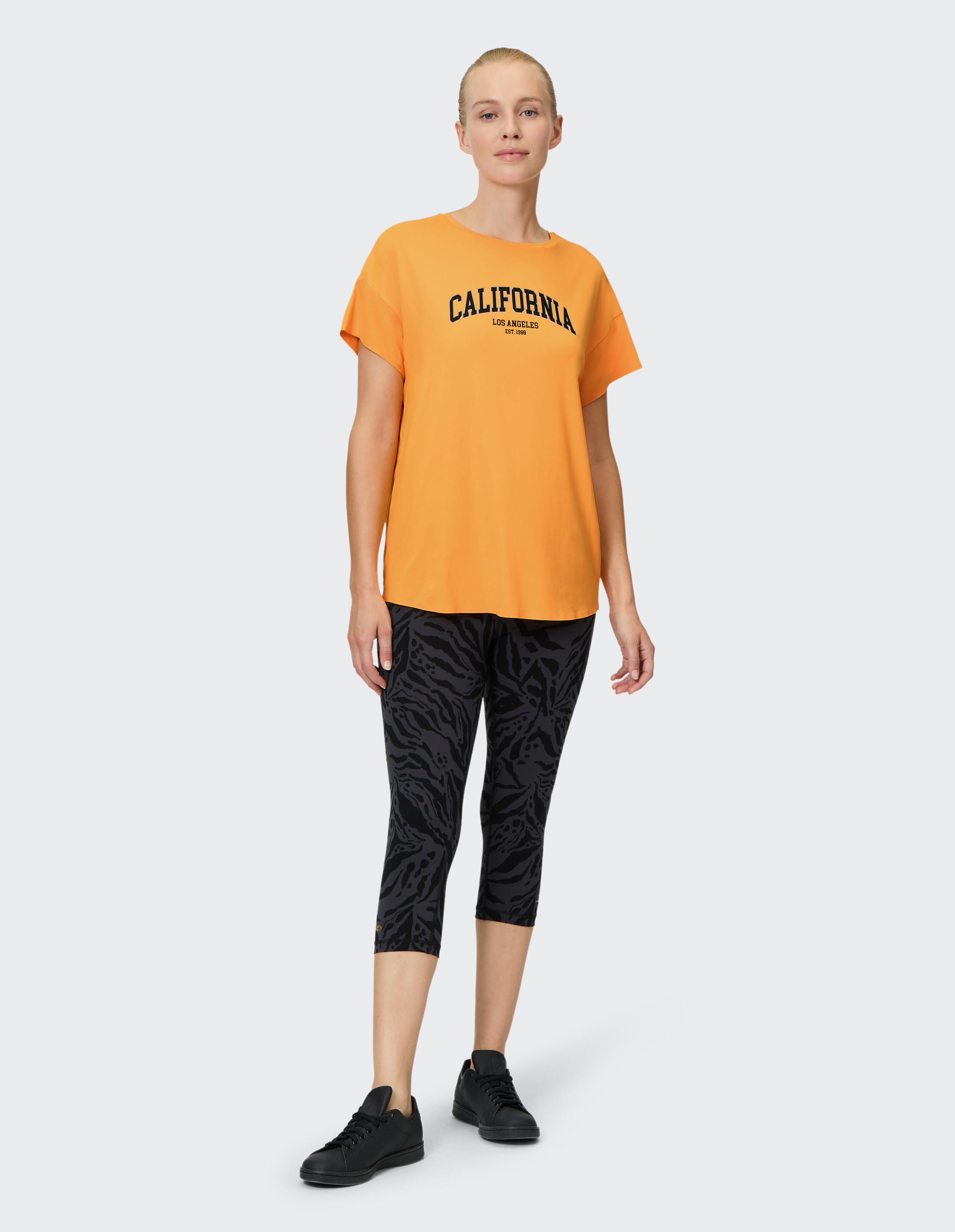 Frau in orangefarbenem California-Grafik-T-Shirt und schwarz gemusterten Leggings, lässiges Outfit.