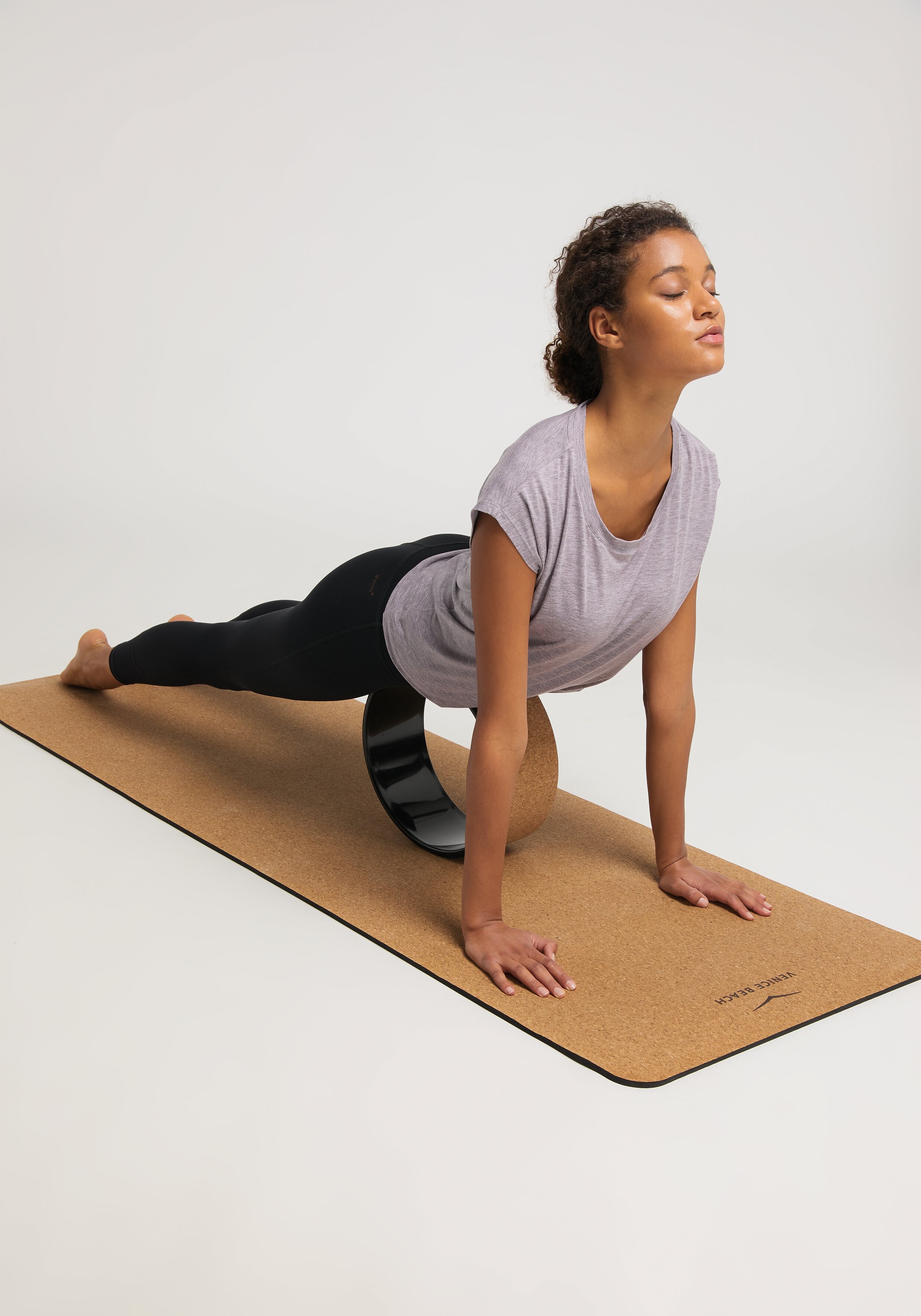 Frau praktiziert Yoga auf einer Korkmatte, trägt ein graues Top und schwarze Leggings.