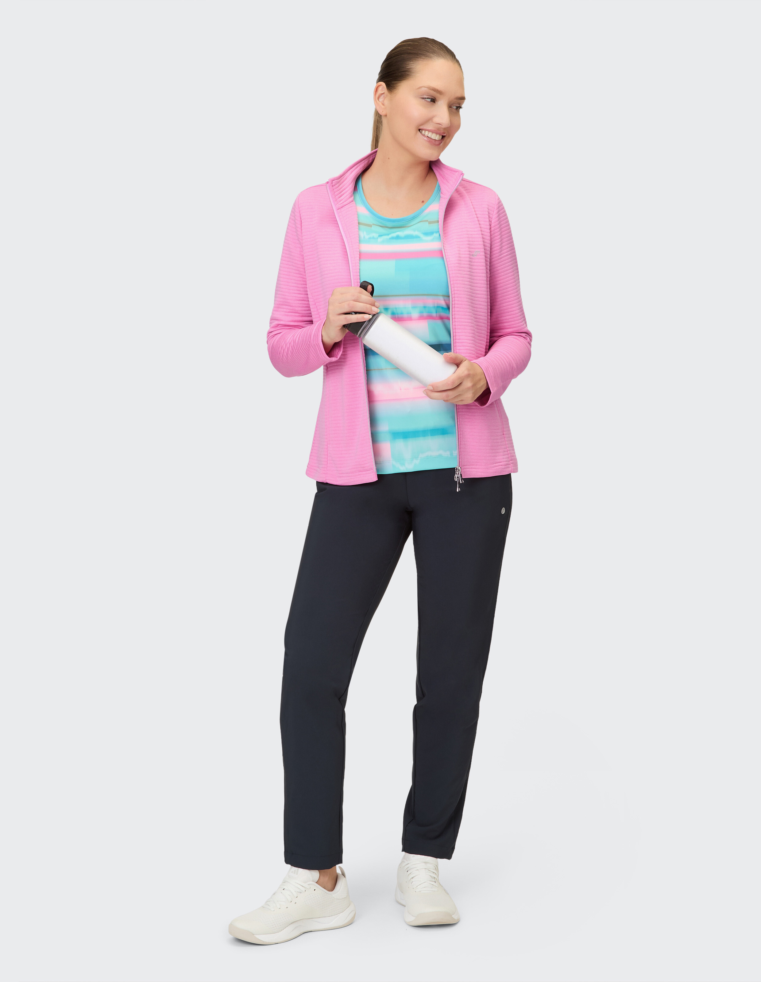 Frau in rosafarbener Jacke mit Reißverschluss und buntem Oberteil, dunkle Hose und Turnschuhe.