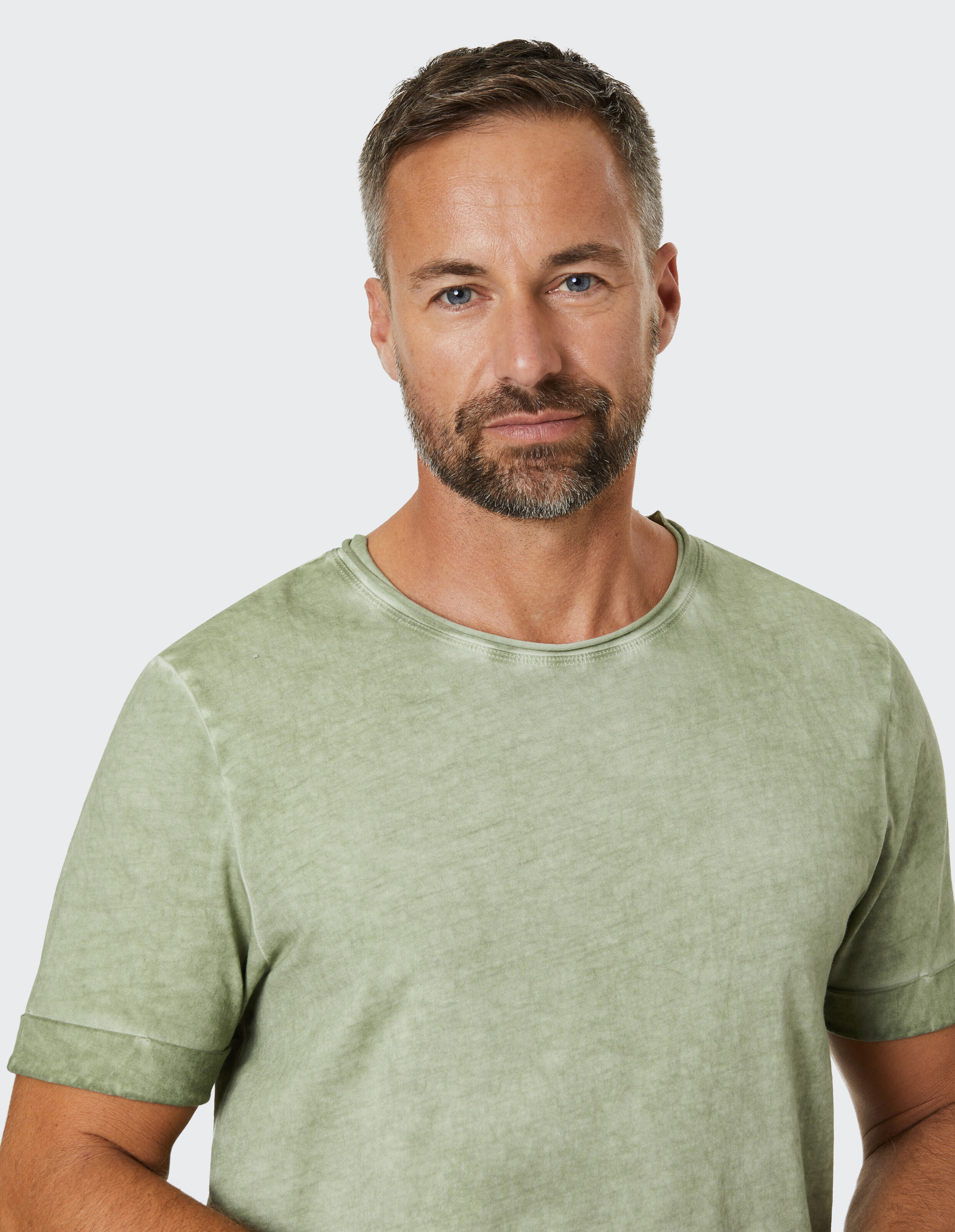 Ein Mann mit kurzem Haar und Bart trägt ein olivgrünes T-Shirt vor einem hellen Hintergrund, schaut direkt in die Kamera.