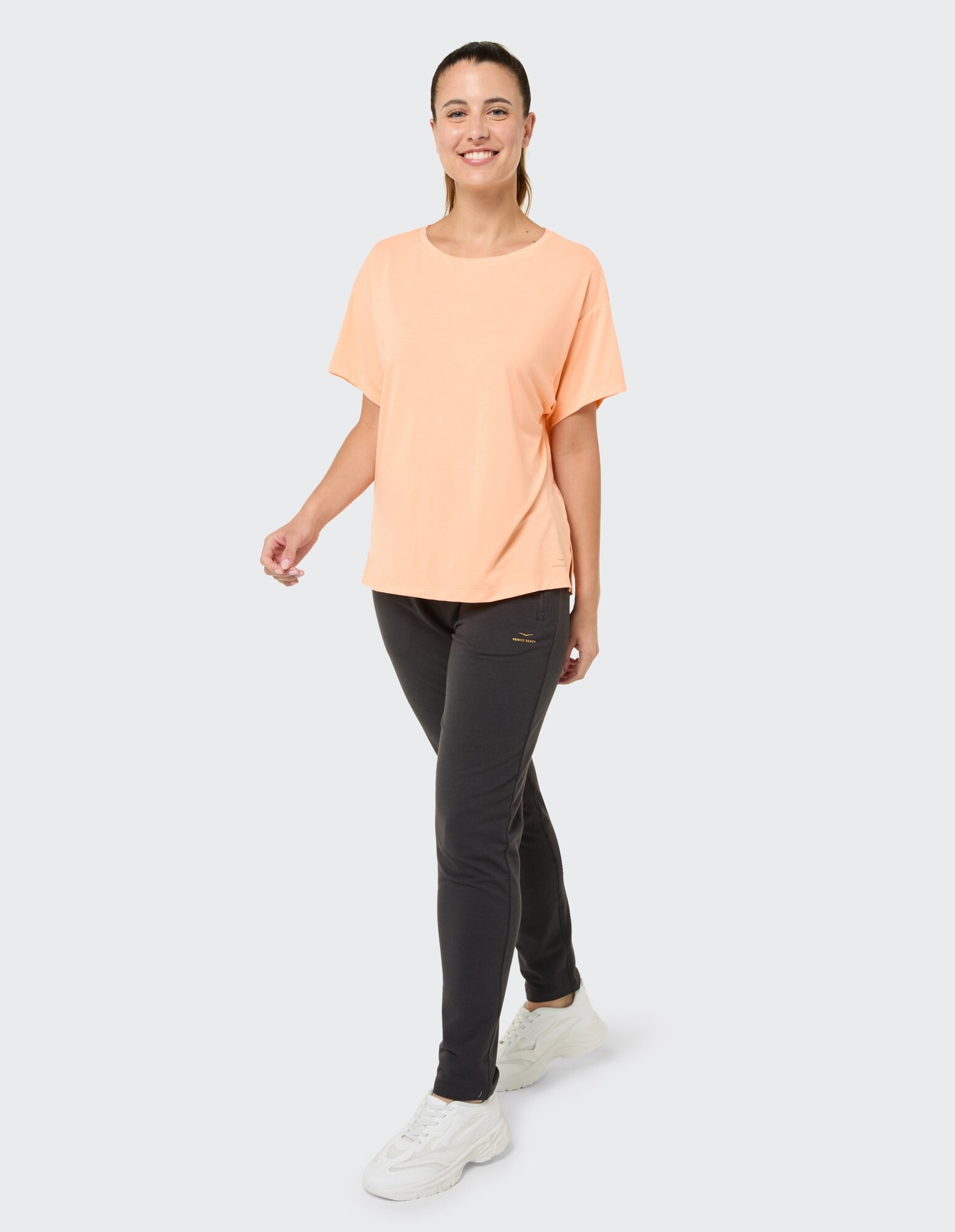 Frau in pfirsichfarbenem T-Shirt und dunkler Hose präsentiert lässige, bequeme Kleidung.