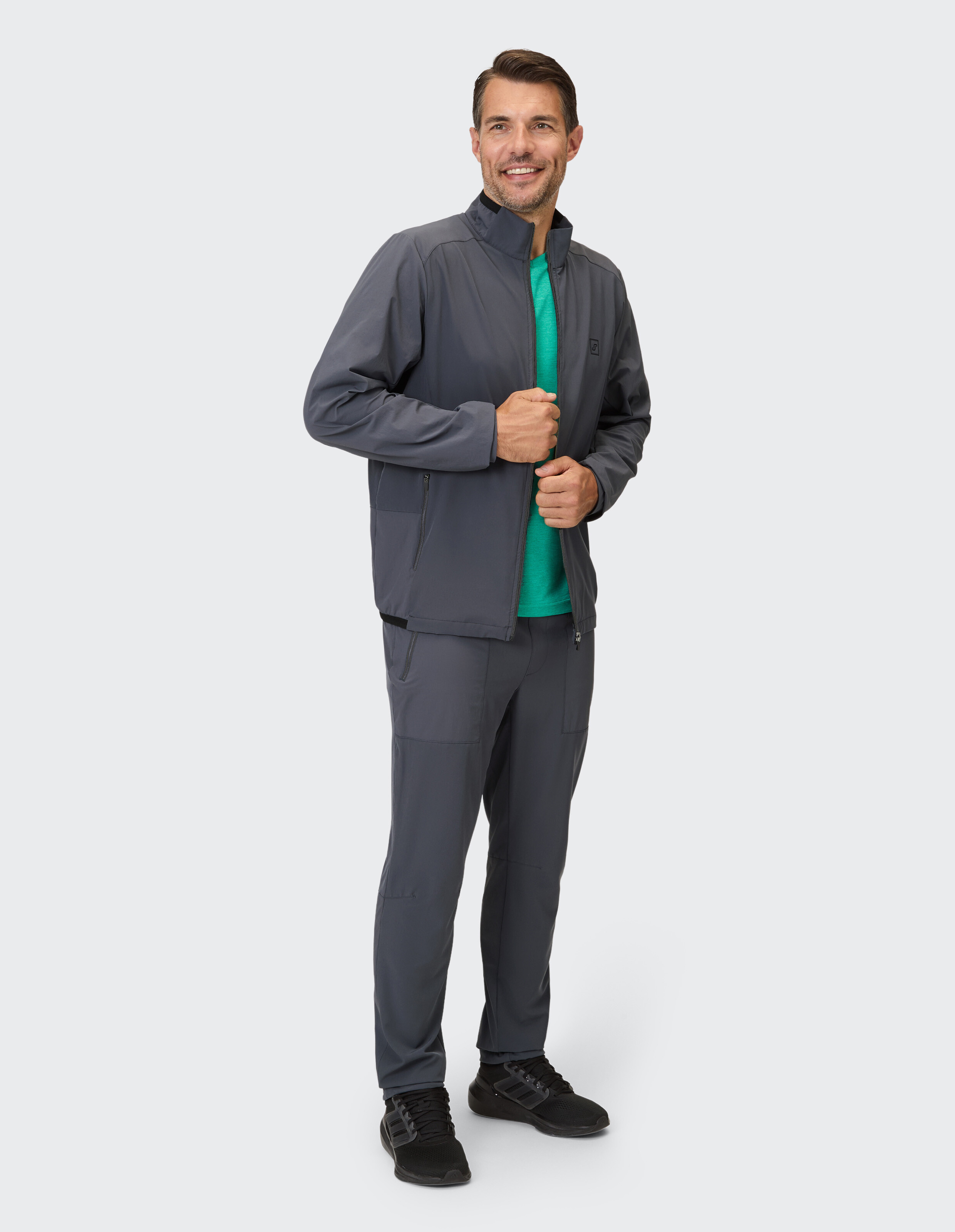 Ein Mann trägt ein dunkelgraues, durchgeknöpftes Jackett mit Reißverschlusstaschen über einem grünen T-Shirt und dazu passende Hose.
