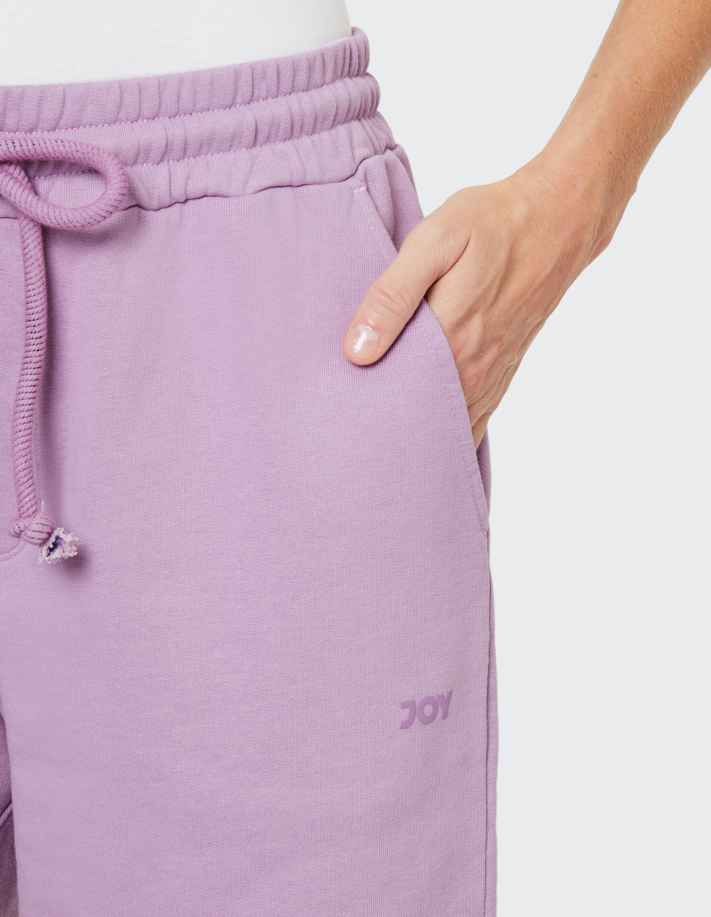 Eine Hand steckt in der Tasche einer lila Jogginghose mit Kordelzug und "JOY"-Aufdruck, getragen von einer Person im hellen Shirt.