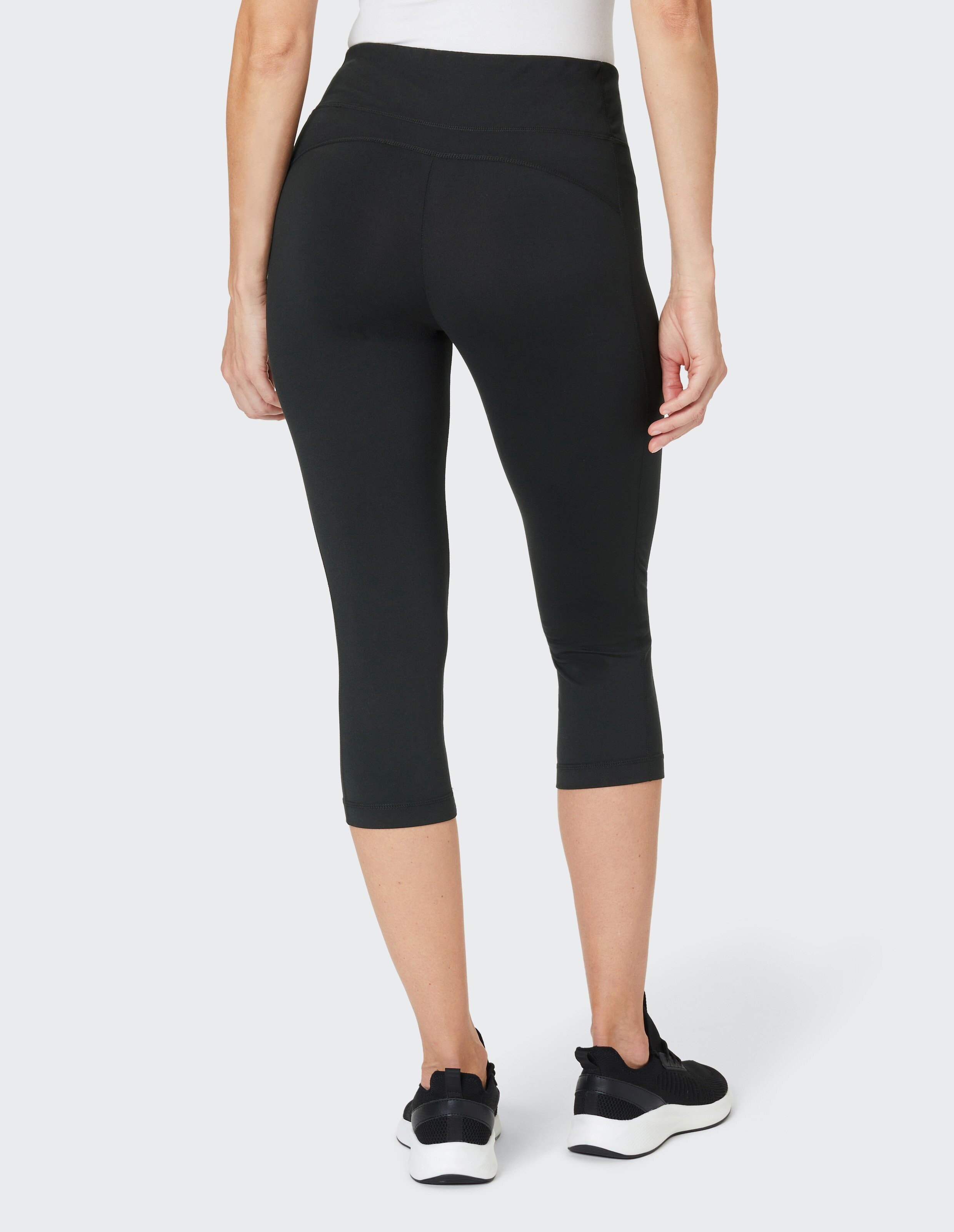 Eine Person trägt schwarze, knielange Leggings und schwarze Sneaker, von hinten fotografiert vor hellem Hintergrund.