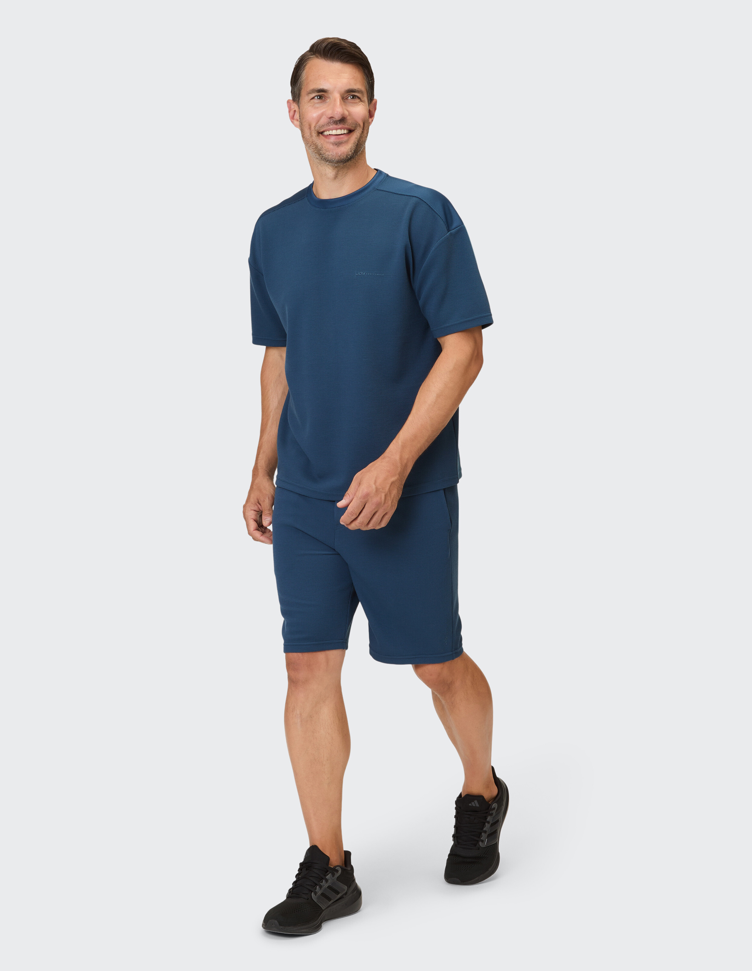Ein Mann in einem dunkelblauen, waffelgemusterten Kurzarmshirt und passenden Shorts, der lächelnd nach rechts blickt.