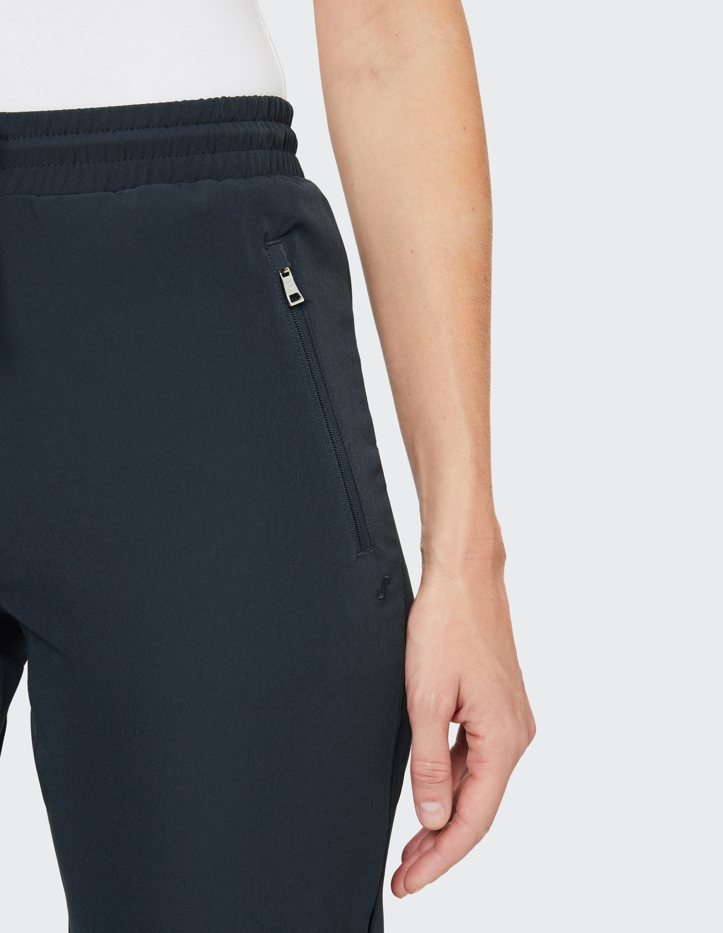 Ein Arm in weißem Oberteil hängt seitlich neben dunkelblauer Hose mit Reißverschlusstasche auf weißem Hintergrund.
