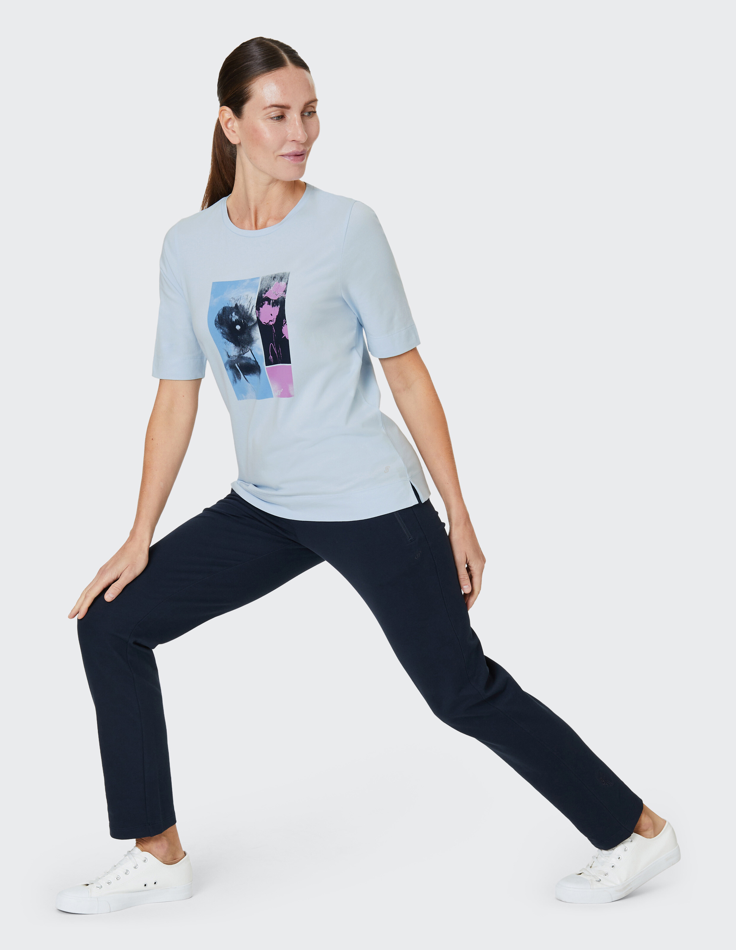 Frau in hellblauem Shirt mit grafischem Druck, dunkler Hose und weißen Sneakern, dehnt sich in seitlicher Ausfallschritt-Pose.