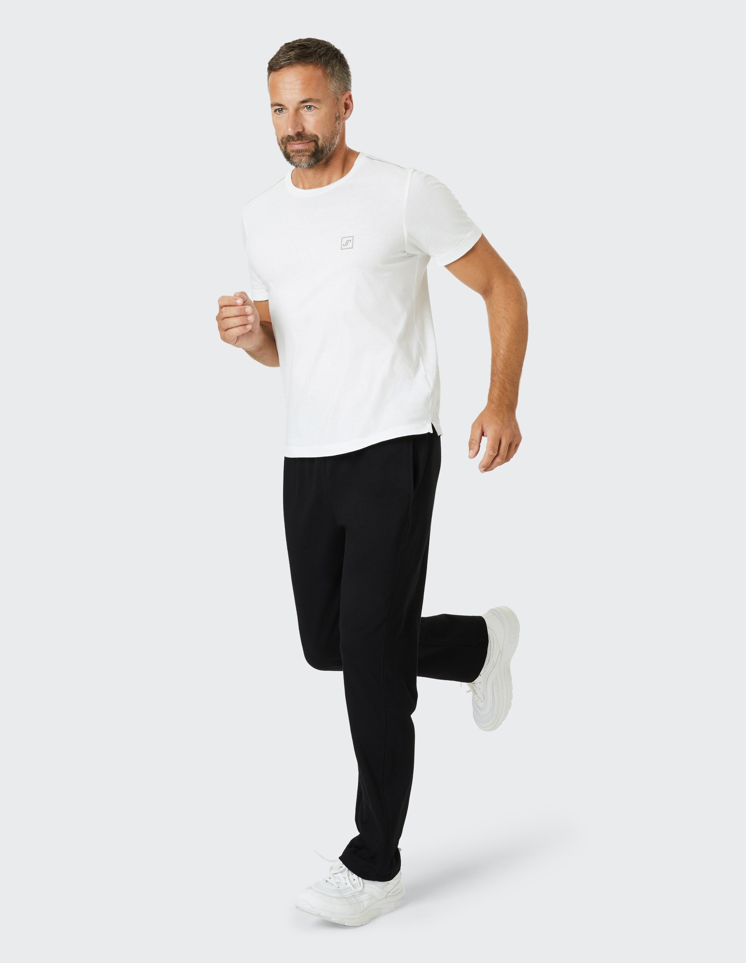 Ein Mann in weißem T-Shirt und schwarzen Jogginghosen läuft auf grauem Hintergrund und trägt weiße Sportschuhe.