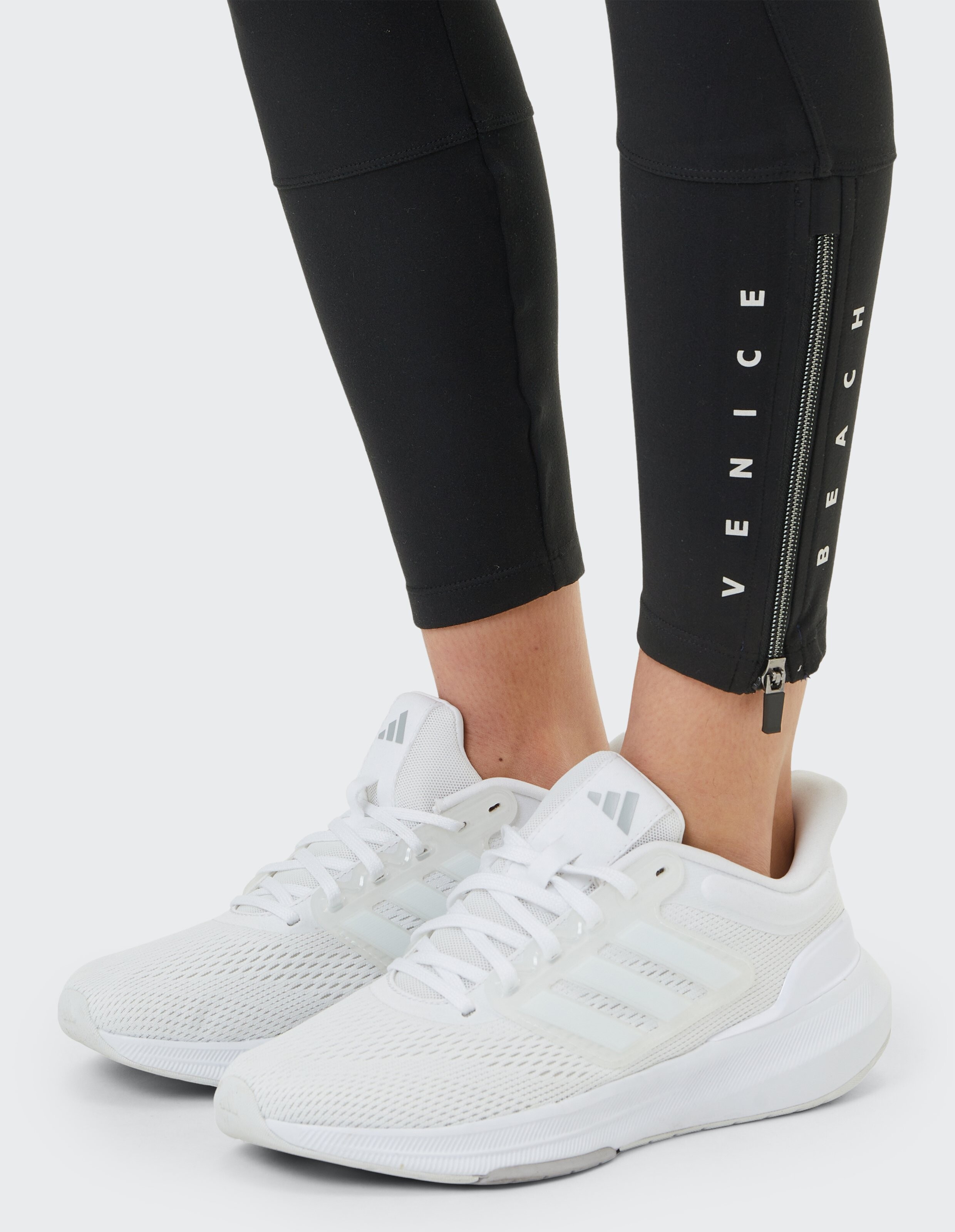 Schwarze Leggings mit „Venice Beach“-Aufdruck und seitlichem Reißverschluss, gepaart mit weißen Sport-Sneakern.