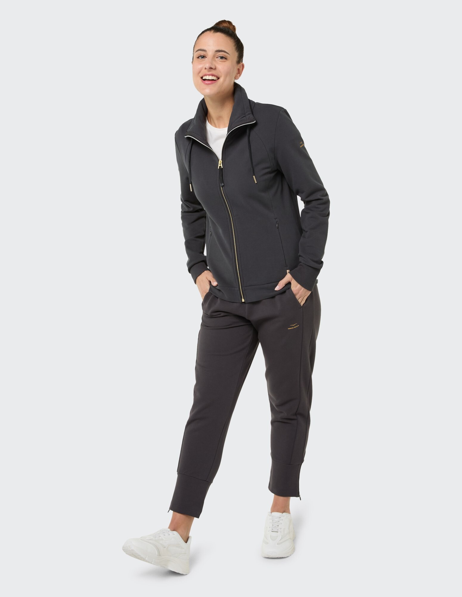 Frau in einer stilvollen grauen Jacke mit Reißverschluss und passender Jogginghose, lächelnd lässig.