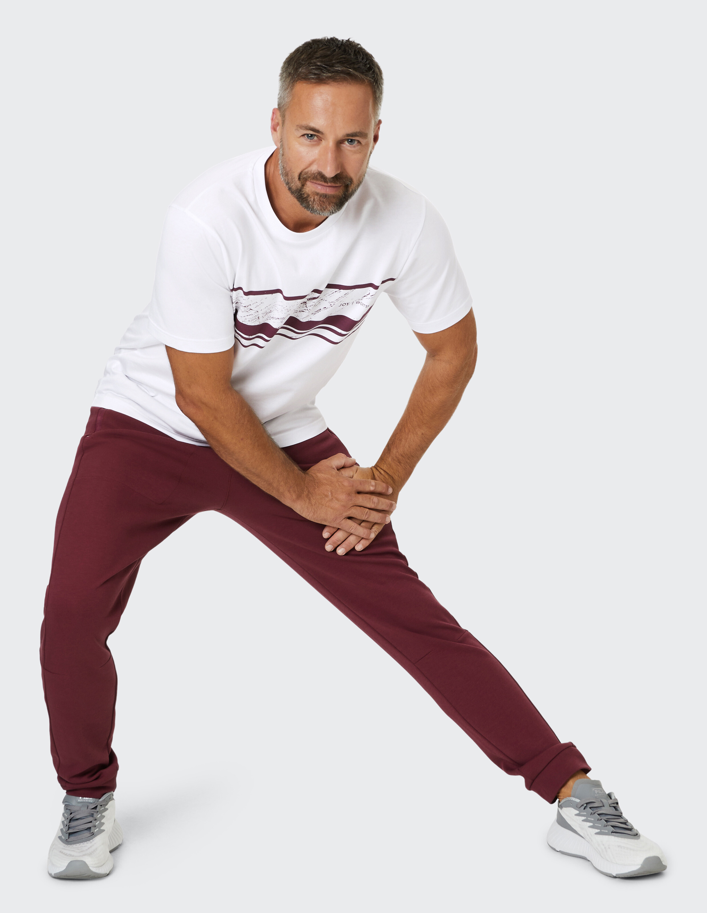 Ein Mann in weißem T-Shirt und bordeauxroten Jogginghosen macht eine Dehnübung auf grauem Hintergrund.