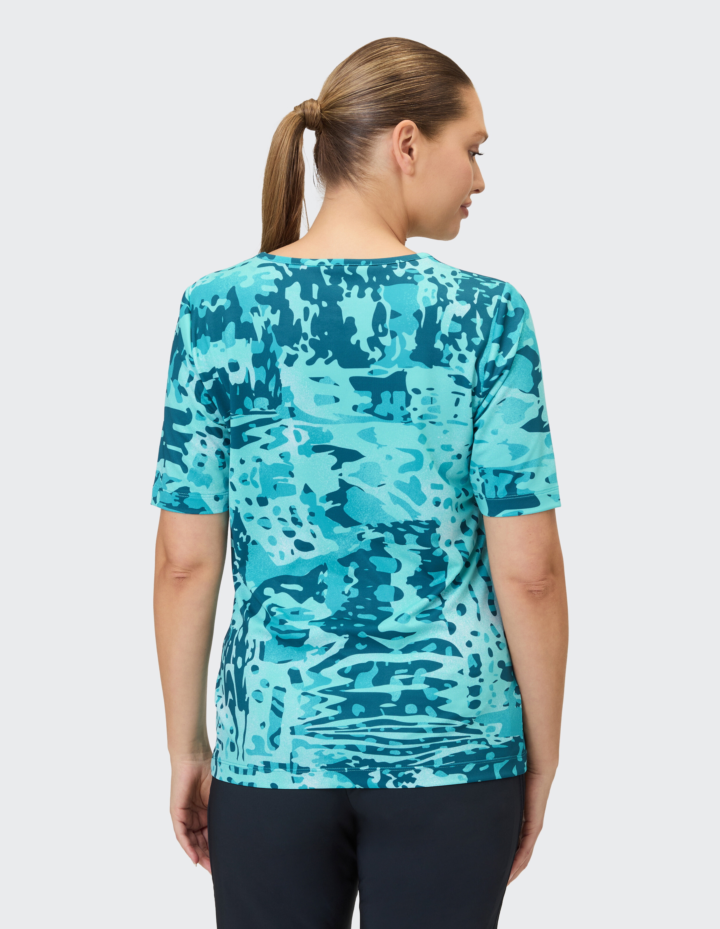Eine Frau mit Pferdeschwanz trägt ein türkisfarbenes T-Shirt mit Camouflage-Muster und kurzärmeligem Schnitt.