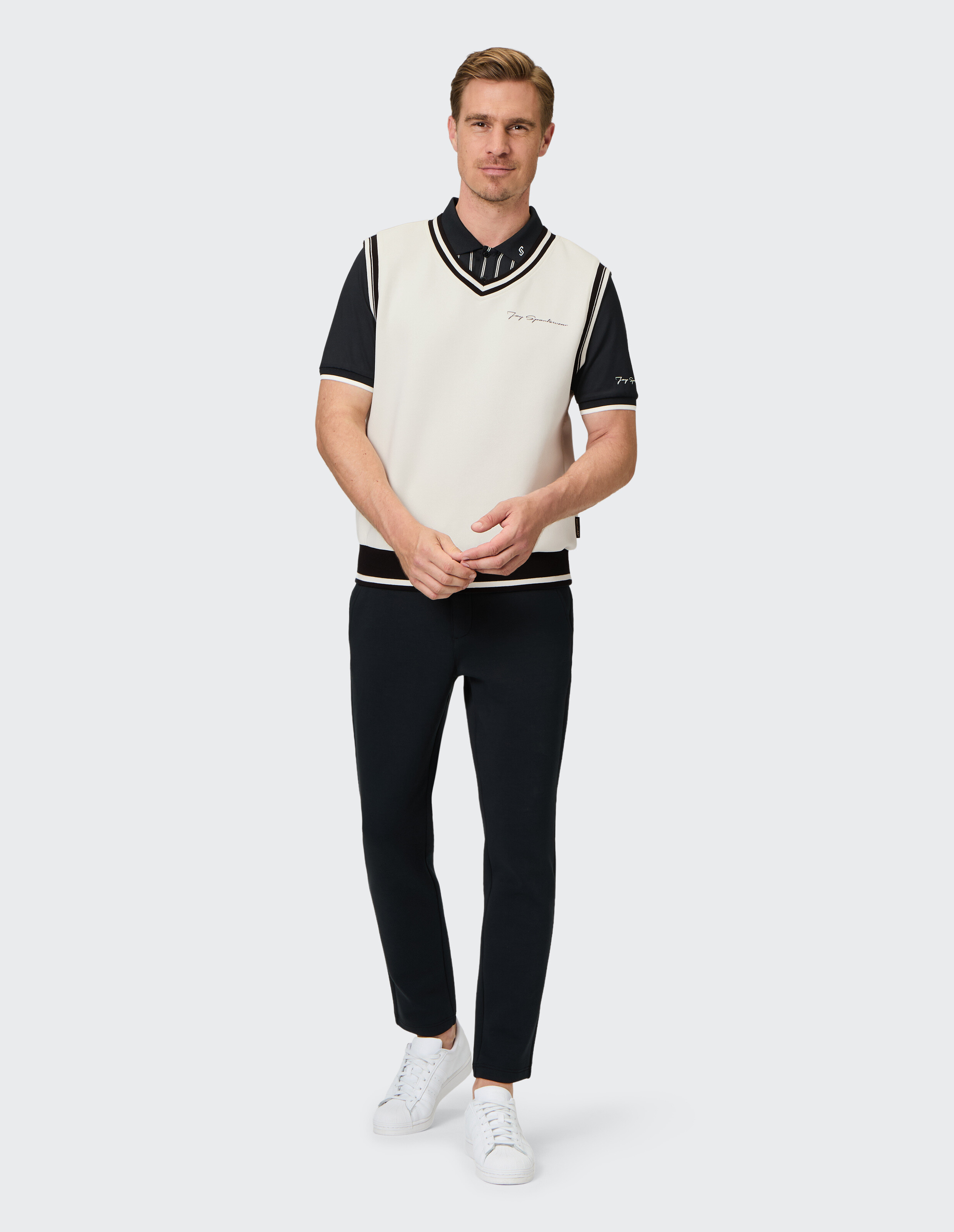 Ein Mann trägt ein schwarz-weißes Outfit mit einem ärmellosen Pullover, darunter ein schwarzes Shirt, schwarze Hosen und weiße Sneaker.