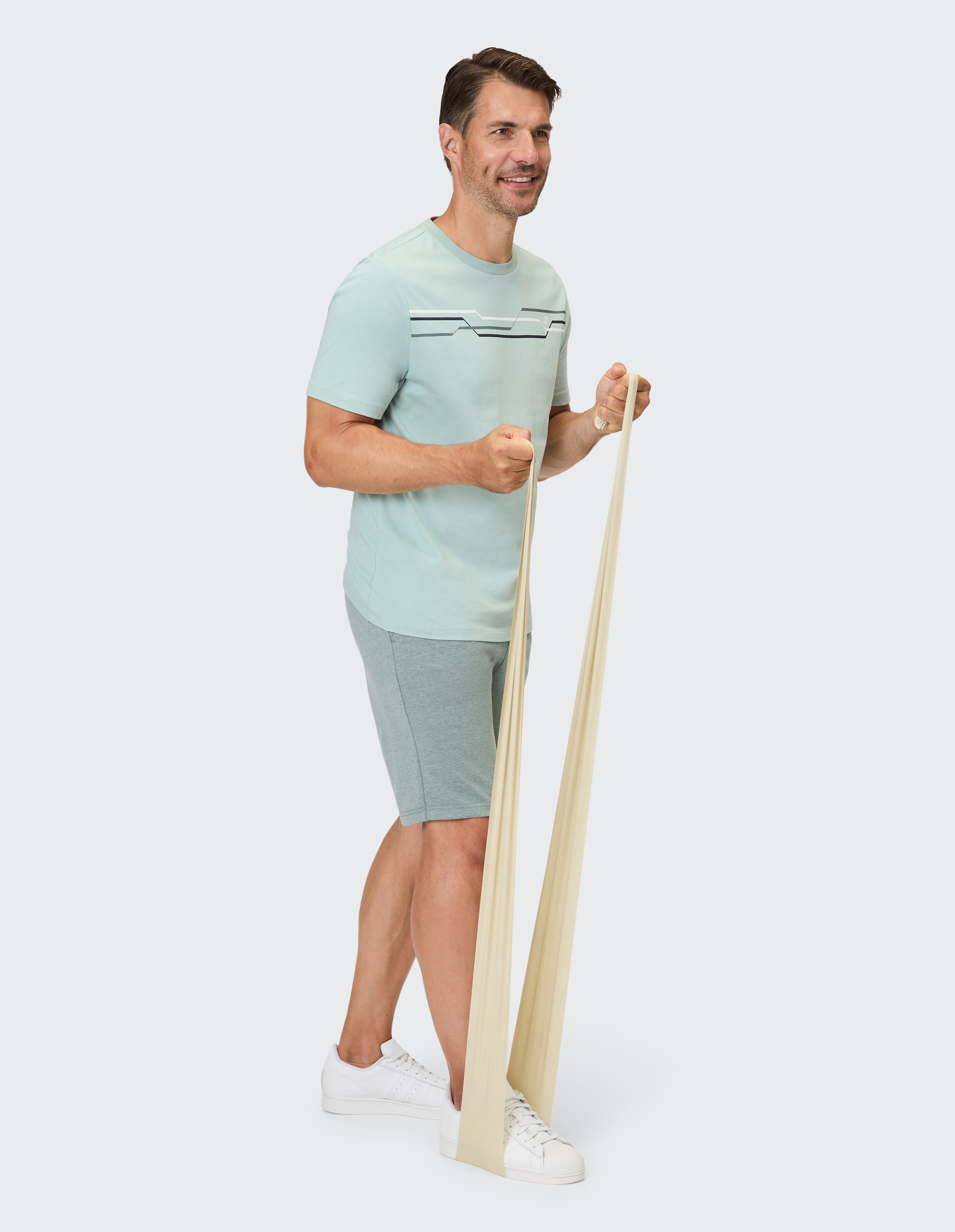 Ein Mann in einem hellgrünen T-Shirt und grauen Shorts trainiert mit einem hellbeigen Widerstandsband.