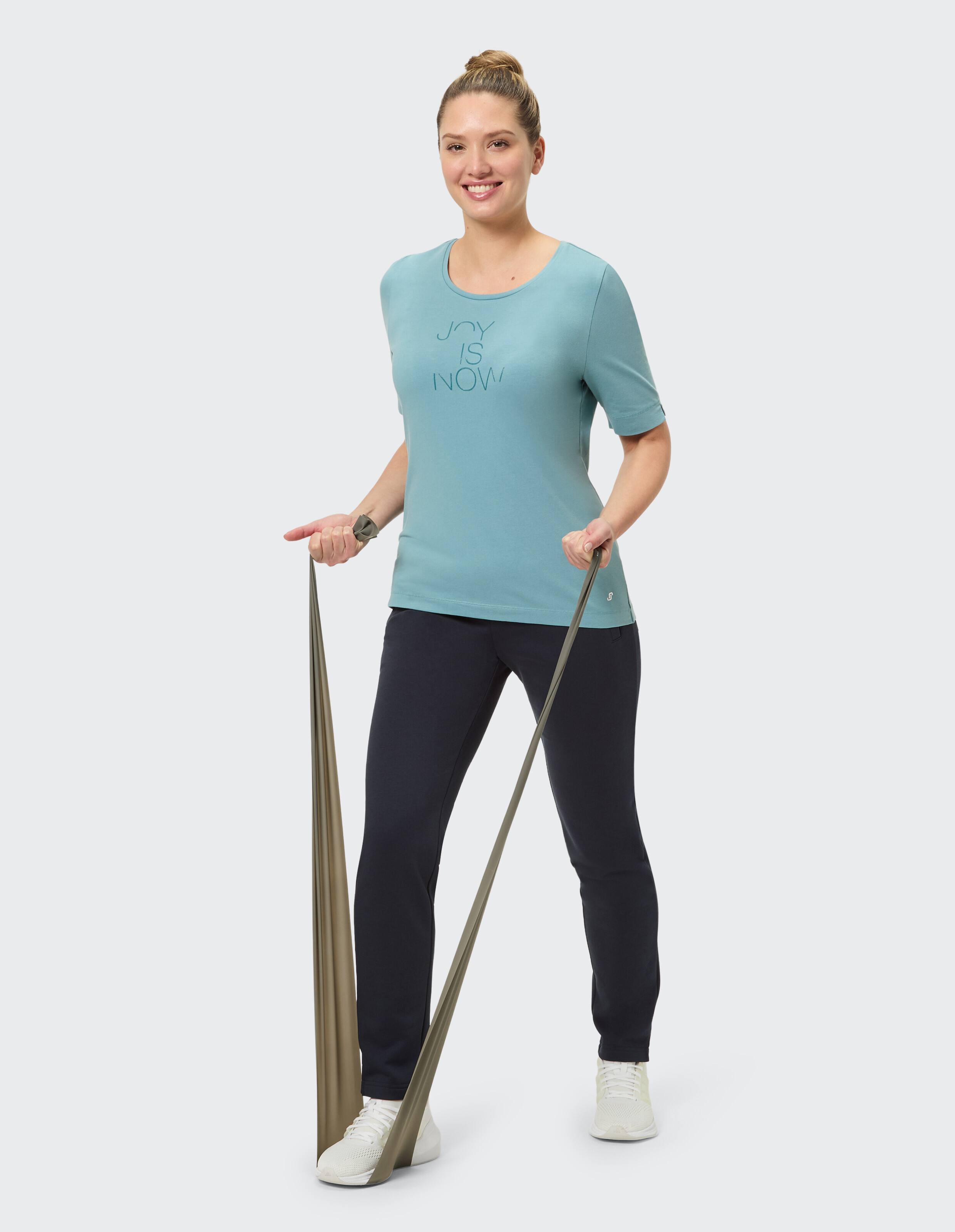 Frau in einem hellblauen T-Shirt mit „Joy is Now“-Aufdruck, dunkler Hose und Turnschuhen.