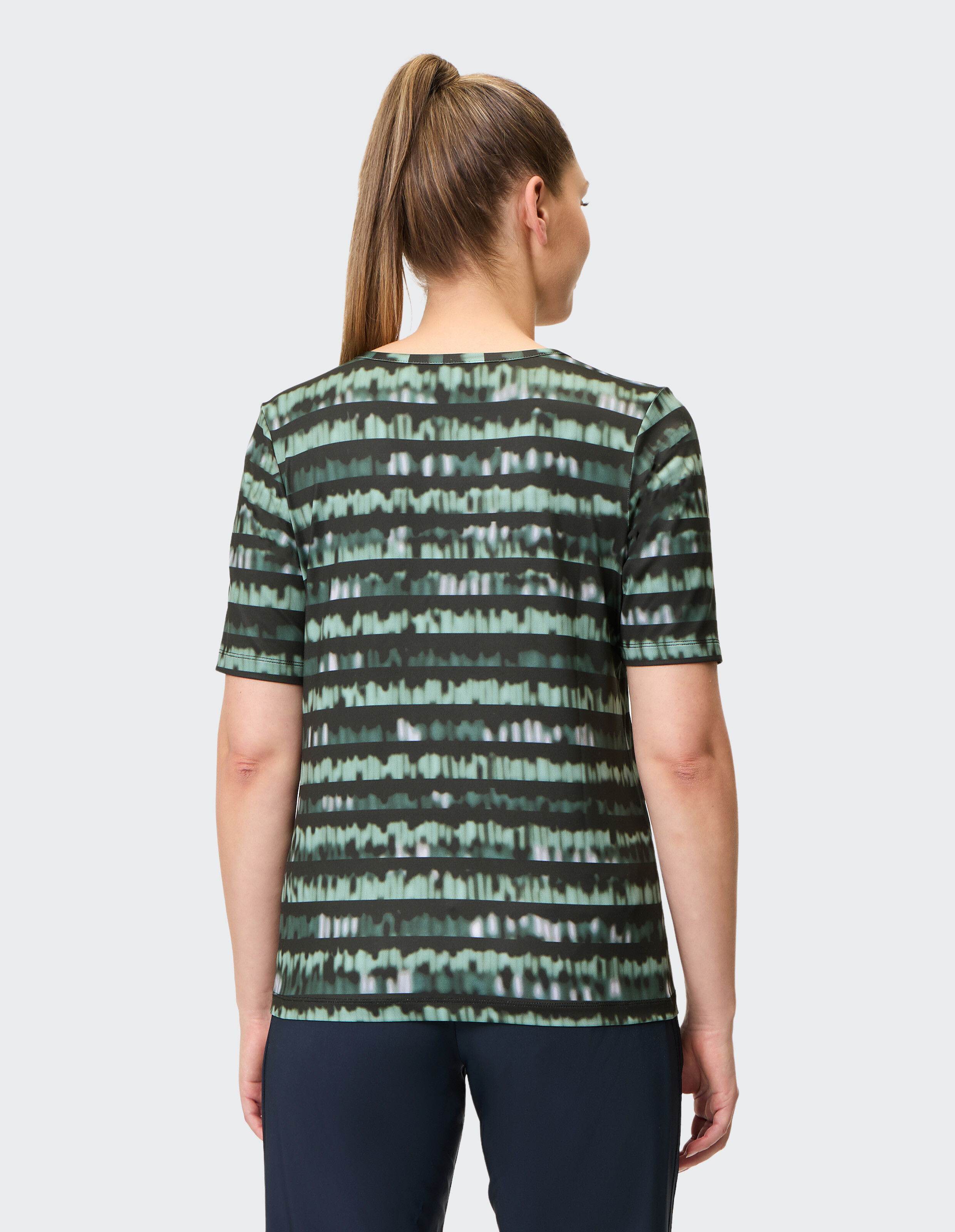 Eine Frau mit langem Pferdeschwanz trägt ein kurzärmeliges, gestreiftes grünes T-Shirt und dunkle Hosen, Rückenansicht.