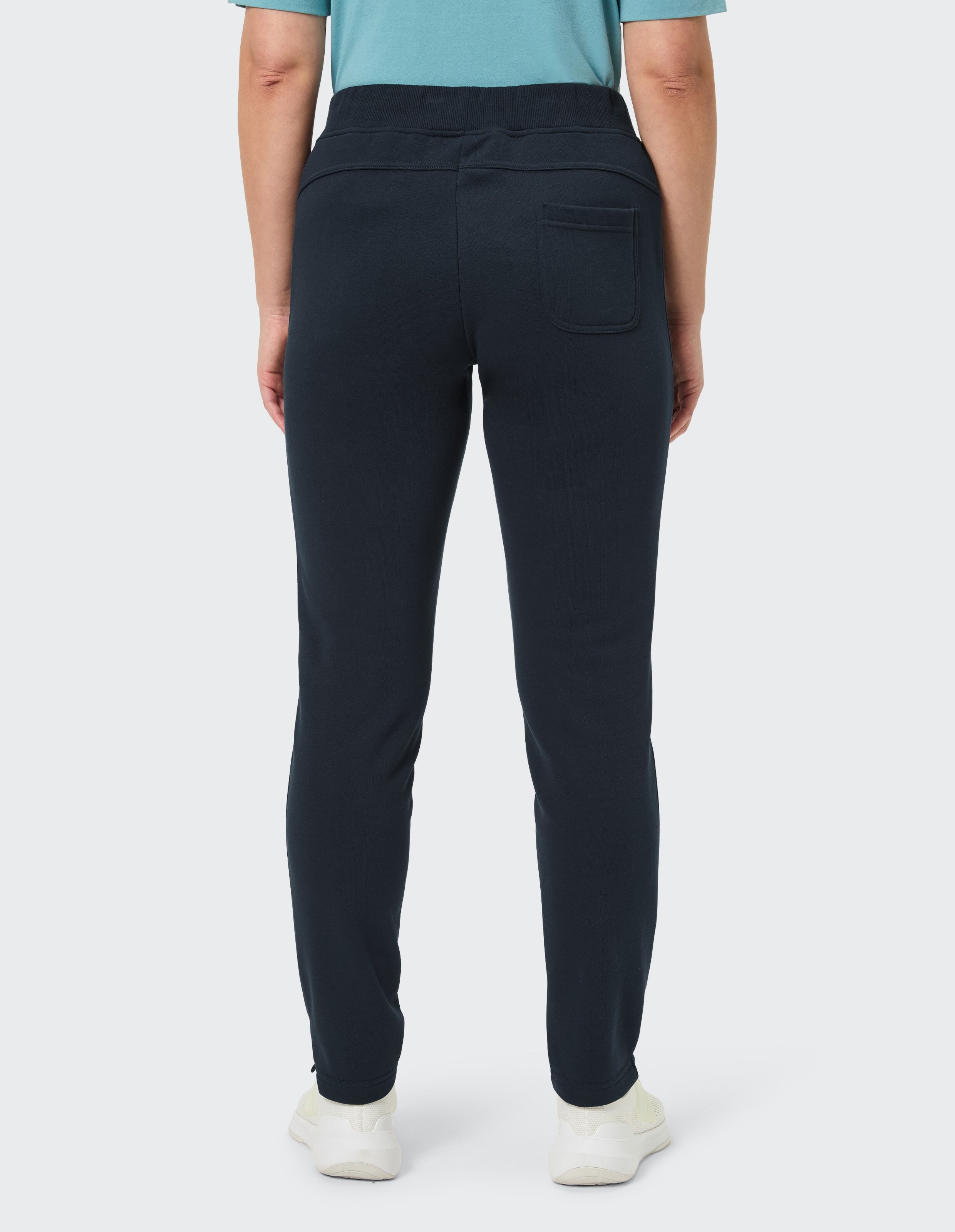 Rückansicht einer Person in einer dunkelblauen Jogginghose mit einer Gesäßtasche, weißem Shirt und rosa Turnschuhen auf weißem Hintergrund.