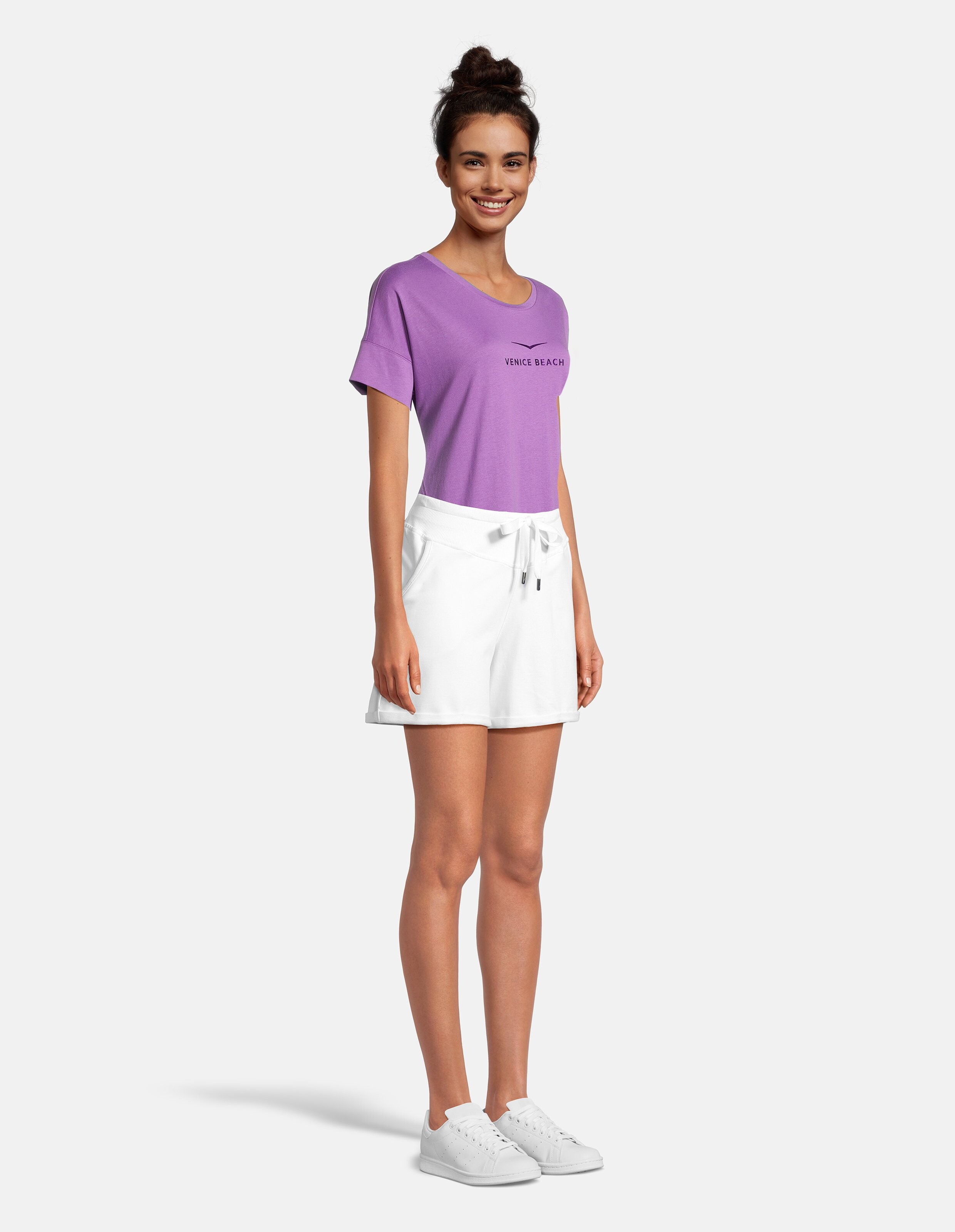 Frau trägt ein lilafarbenes Venice Beach-T-Shirt und weiße Freizeitshorts zum Entspannen.