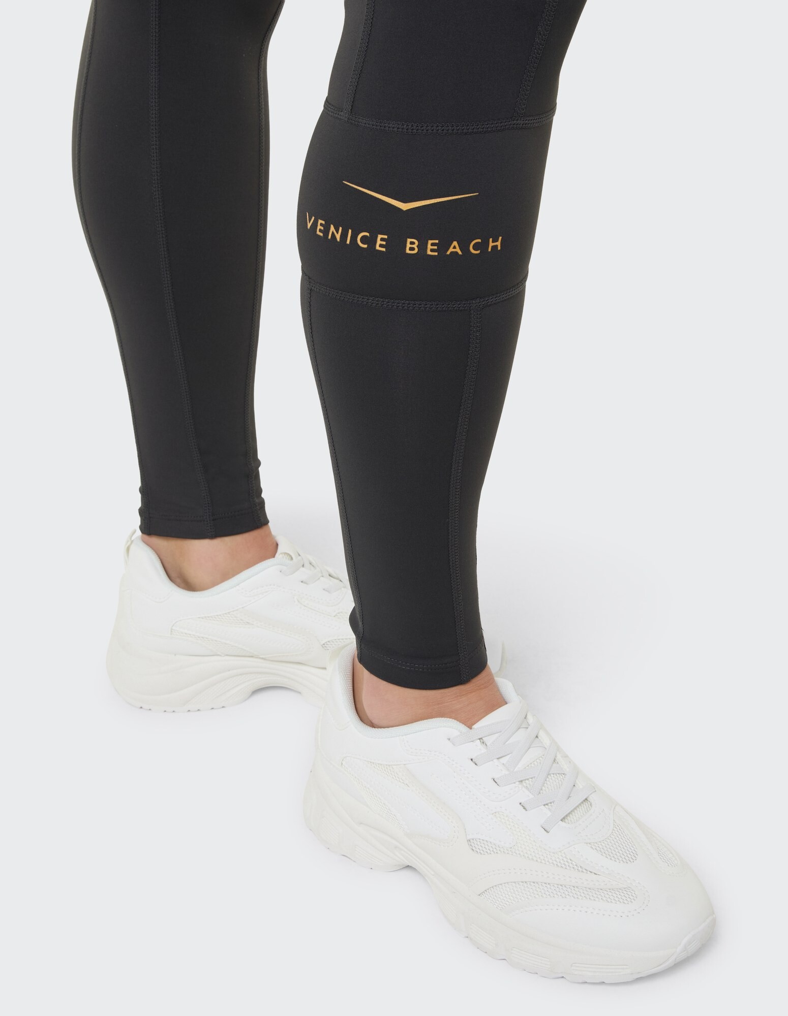 Schwarze Leggings mit goldenem „Venice Beach“-Logo, gepaart mit weißen Sneakers.