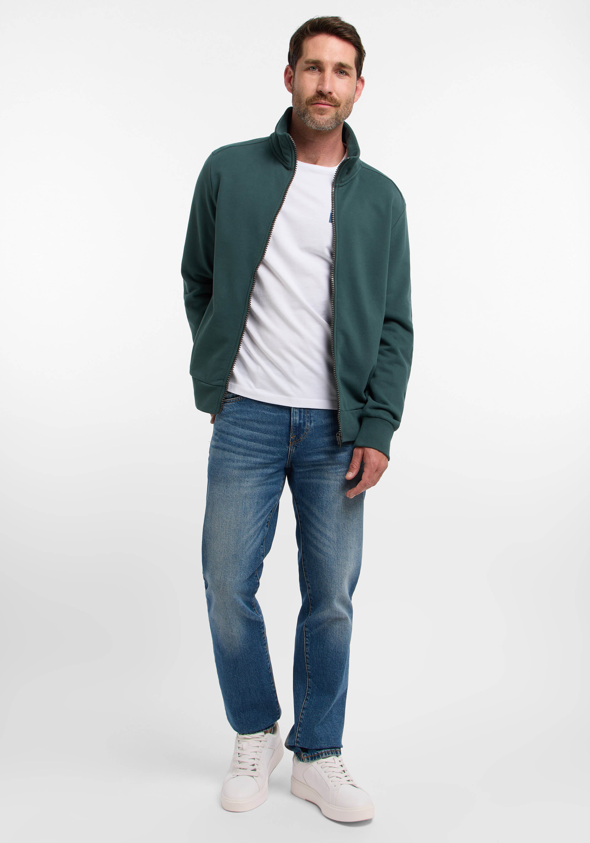 Mann in einer grünen Jacke mit Reißverschluss und blauen Jeans, gestylt für Freizeitkleidung.