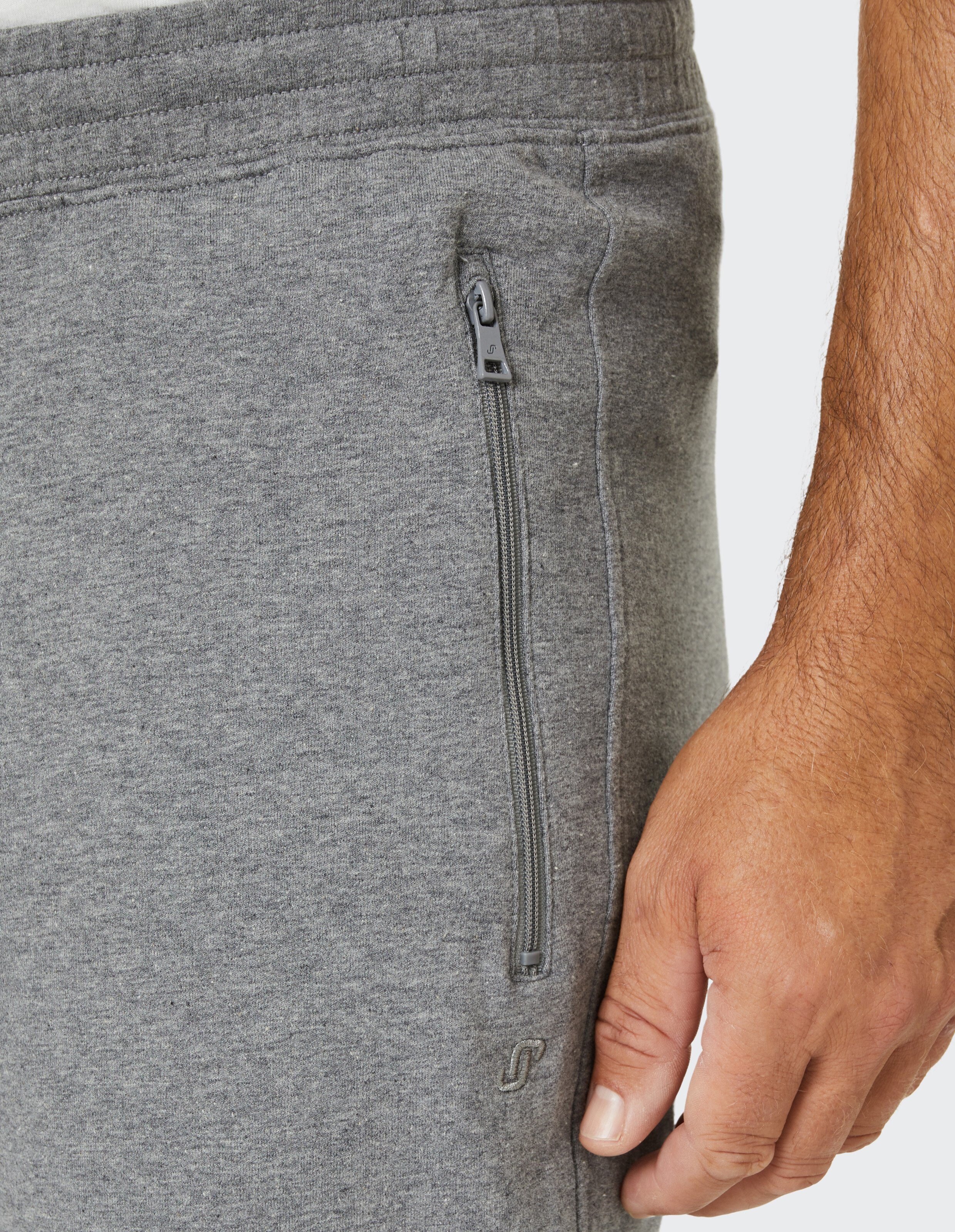 Graue Jogginghose mit seitlichem Reißverschluss und leicht sichtbarem Logo, getragen von einer Person, deren Hand zu sehen ist.