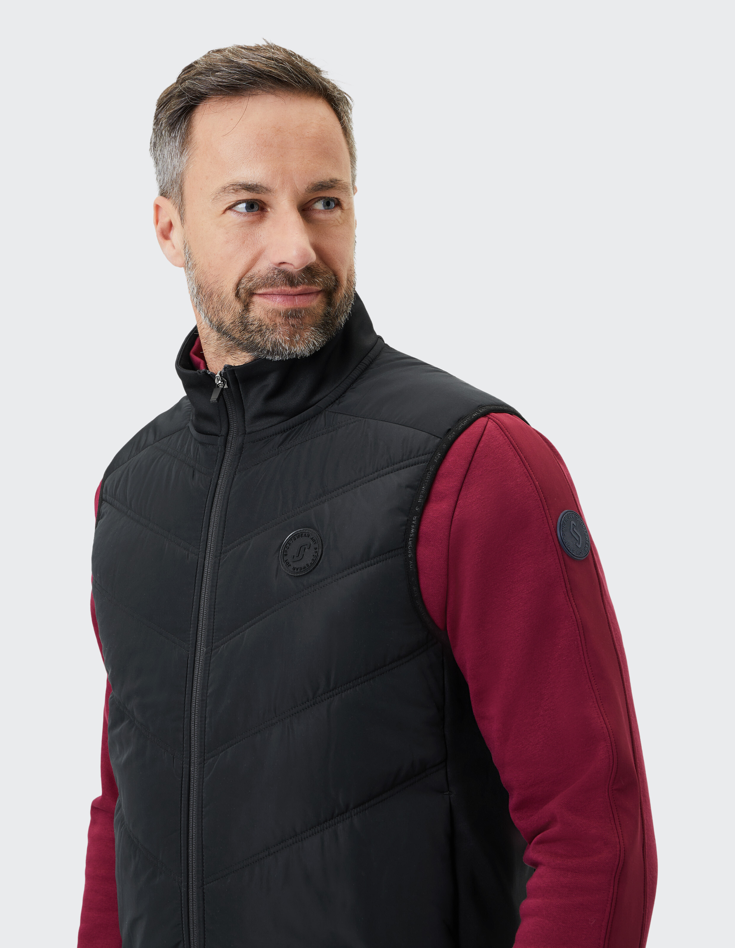 Mann trägt eine schwarze gesteppte ELBSAND-Weste über einem burgunderroten Freizeitpullover.
