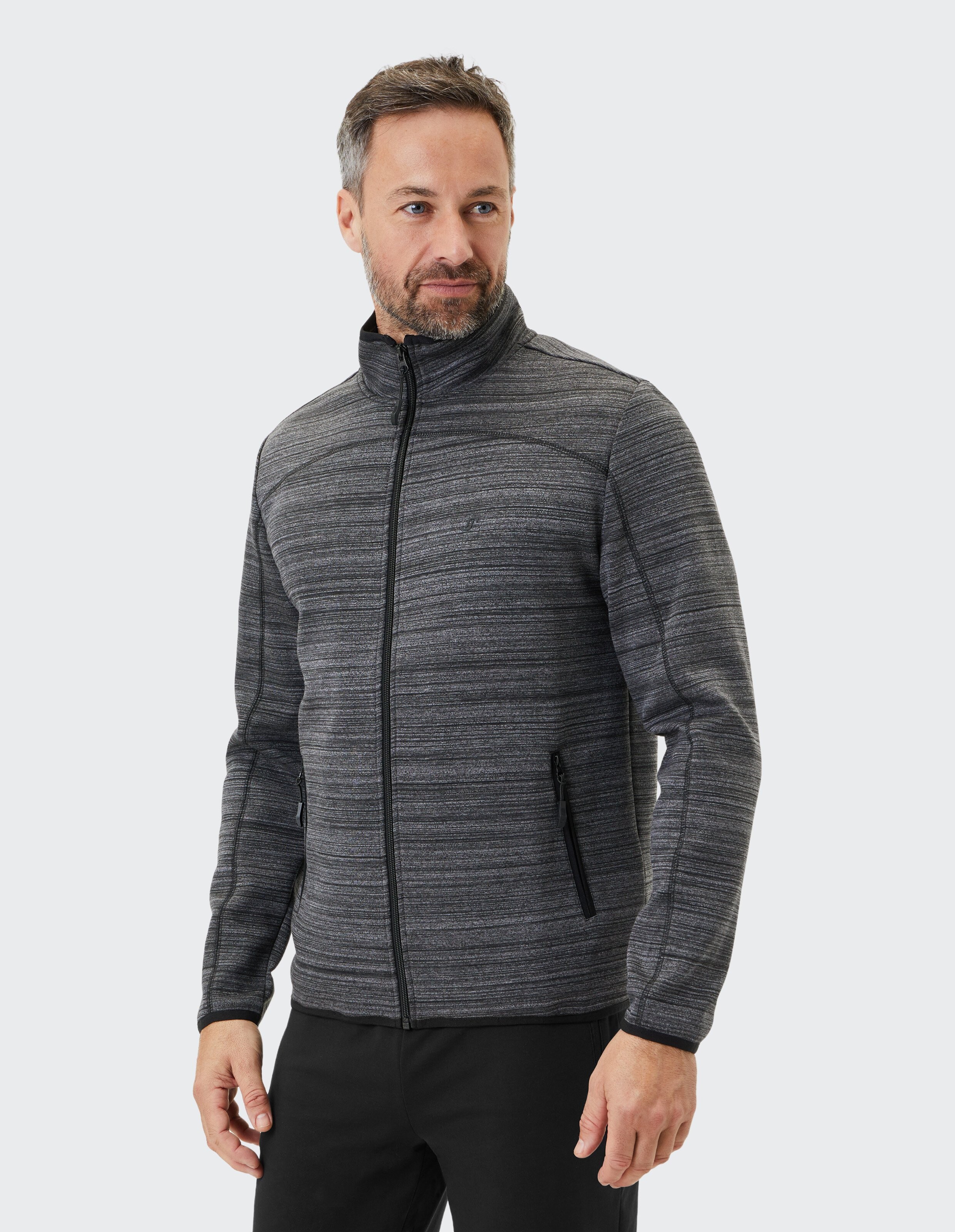 Ein lächelnder Mann trägt eine dunkelgraue, gestreifte Jacke mit Reißverschluss und Stehkragen vor einem hellen Hintergrund.