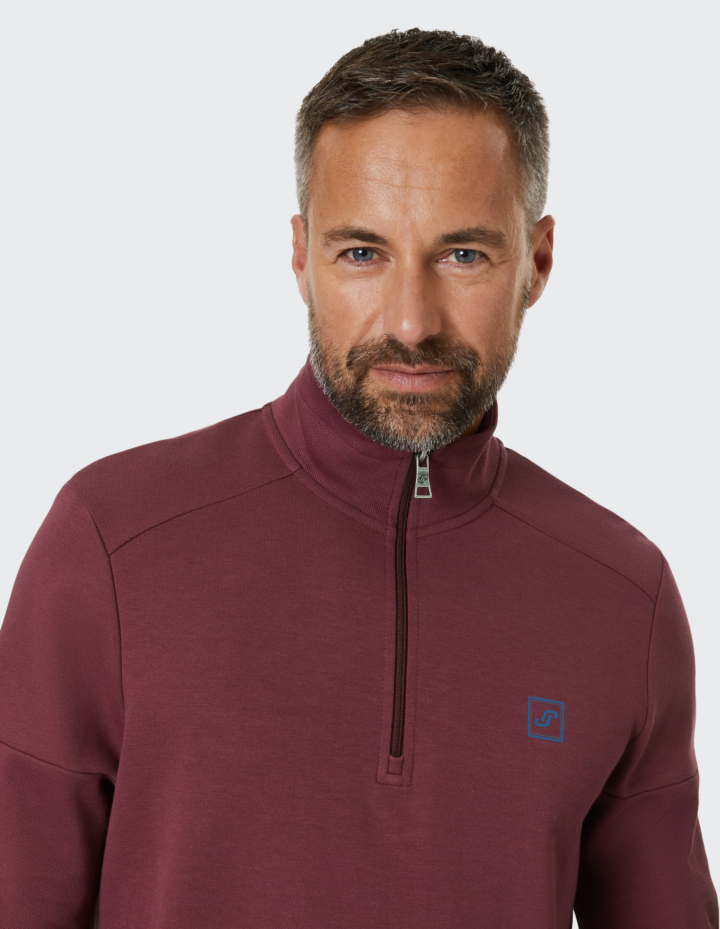 Ein Mann mit kurzem Haar und Bart trägt einen weinroten Pullover mit halb geöffnetem Reißverschluss vor einem hellgrauen Hintergrund.