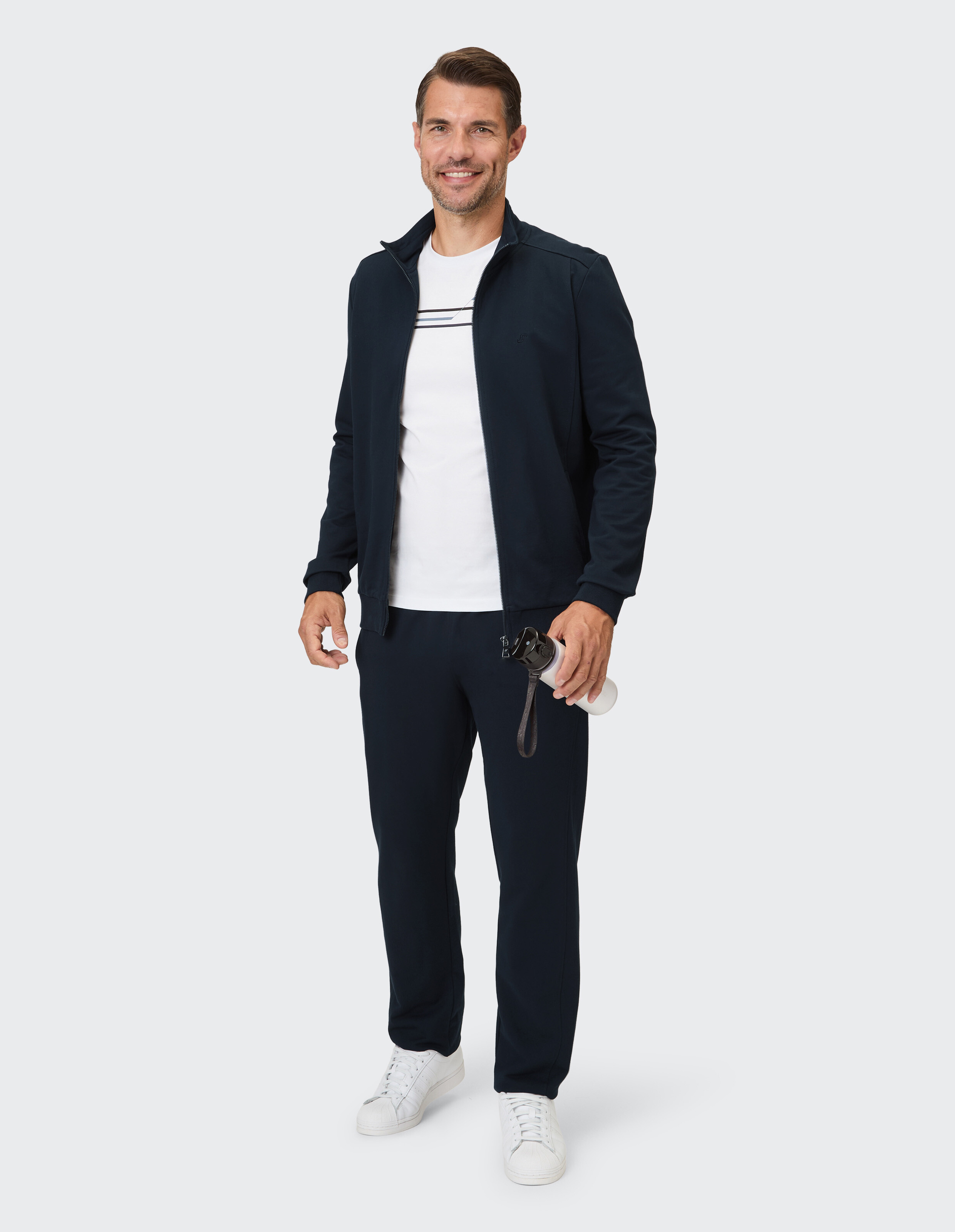 Ein Mann trägt einen dunkelblauen Trainingsanzug bestehend aus einer Jacke mit durchgehendem Reißverschluss und einer Hose, dazu ein weißes T-Shirt mit Streifen.