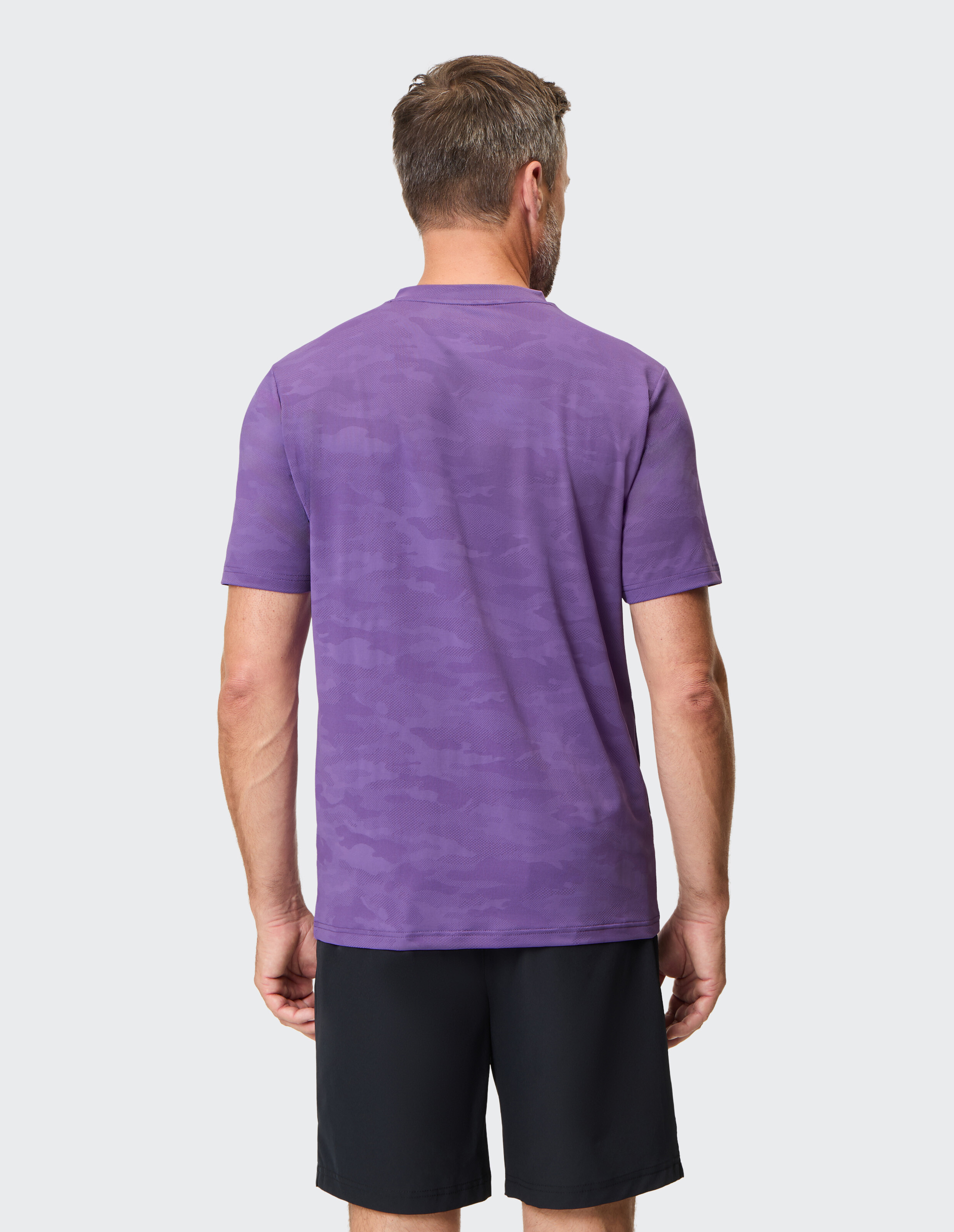 Mann trägt ein lilafarbenes Freizeit-T-Shirt mit Tarnmuster und schwarze Shorts, Rückansicht.