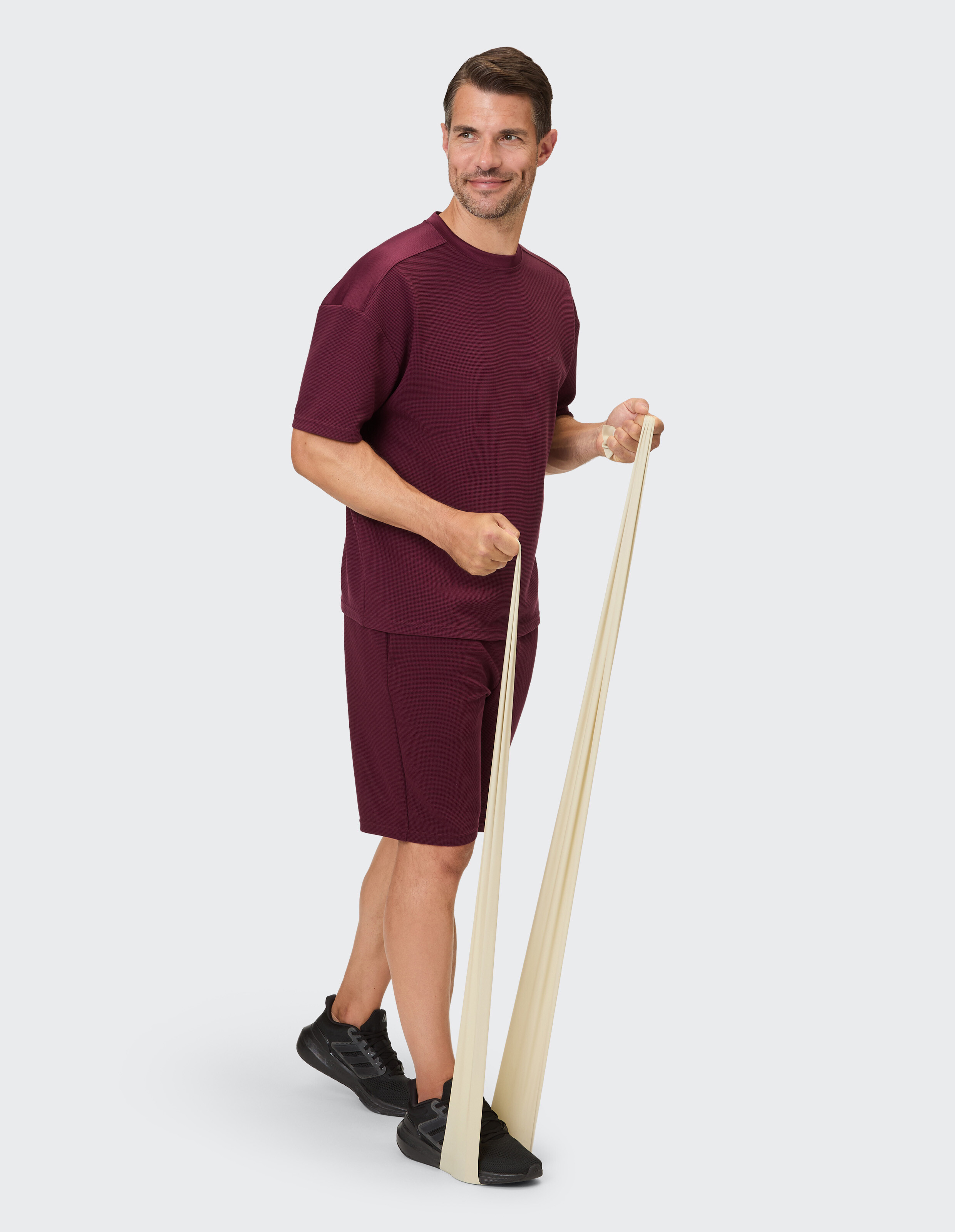 Ein Mann in bordeauxrotem Trainingsset trainiert mit einem hellbeigen Widerstandsband.