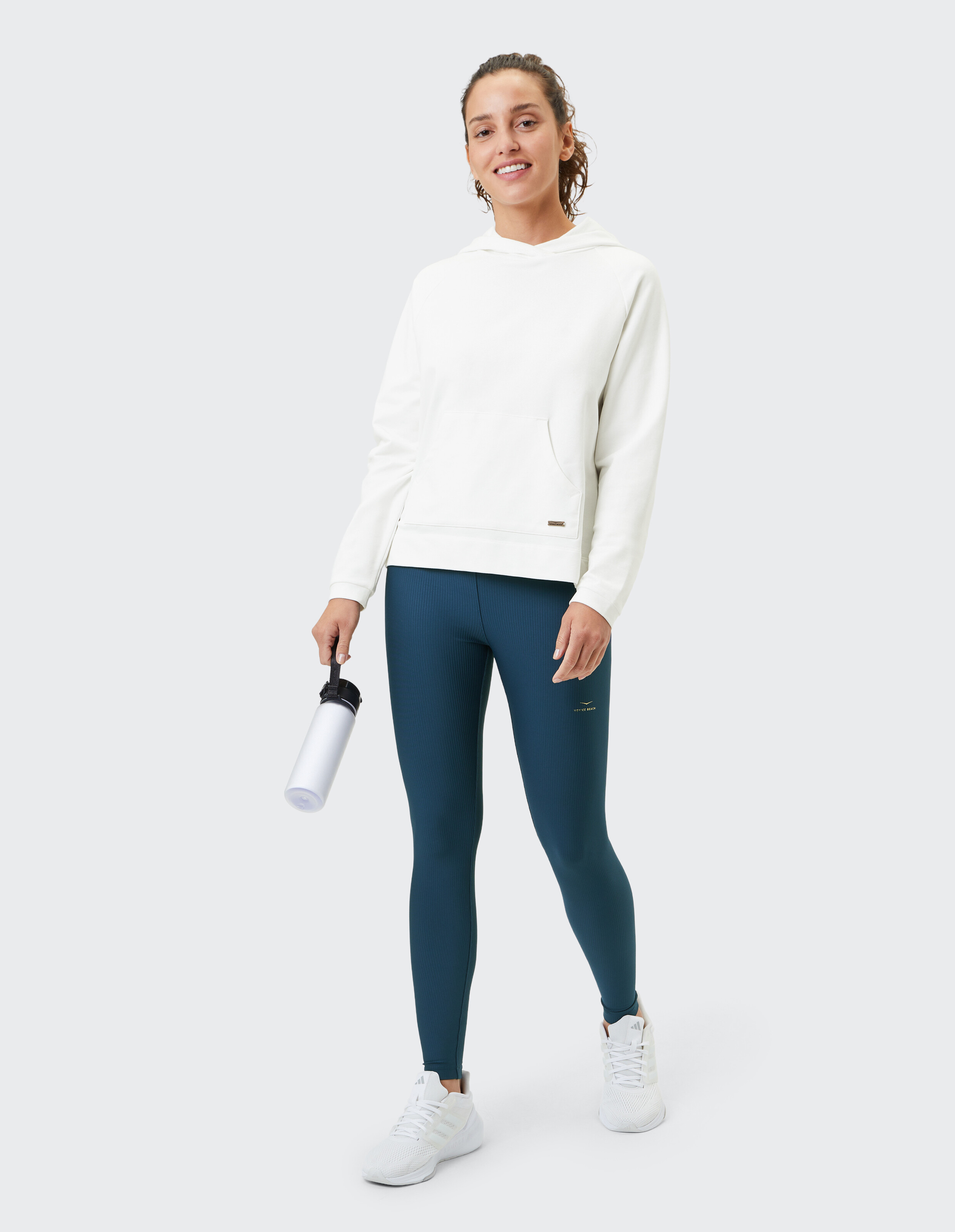 Frau in einem weißen Kapuzenpullover und blauen Leggings mit einer Wasserflasche für einen lässigen Look.