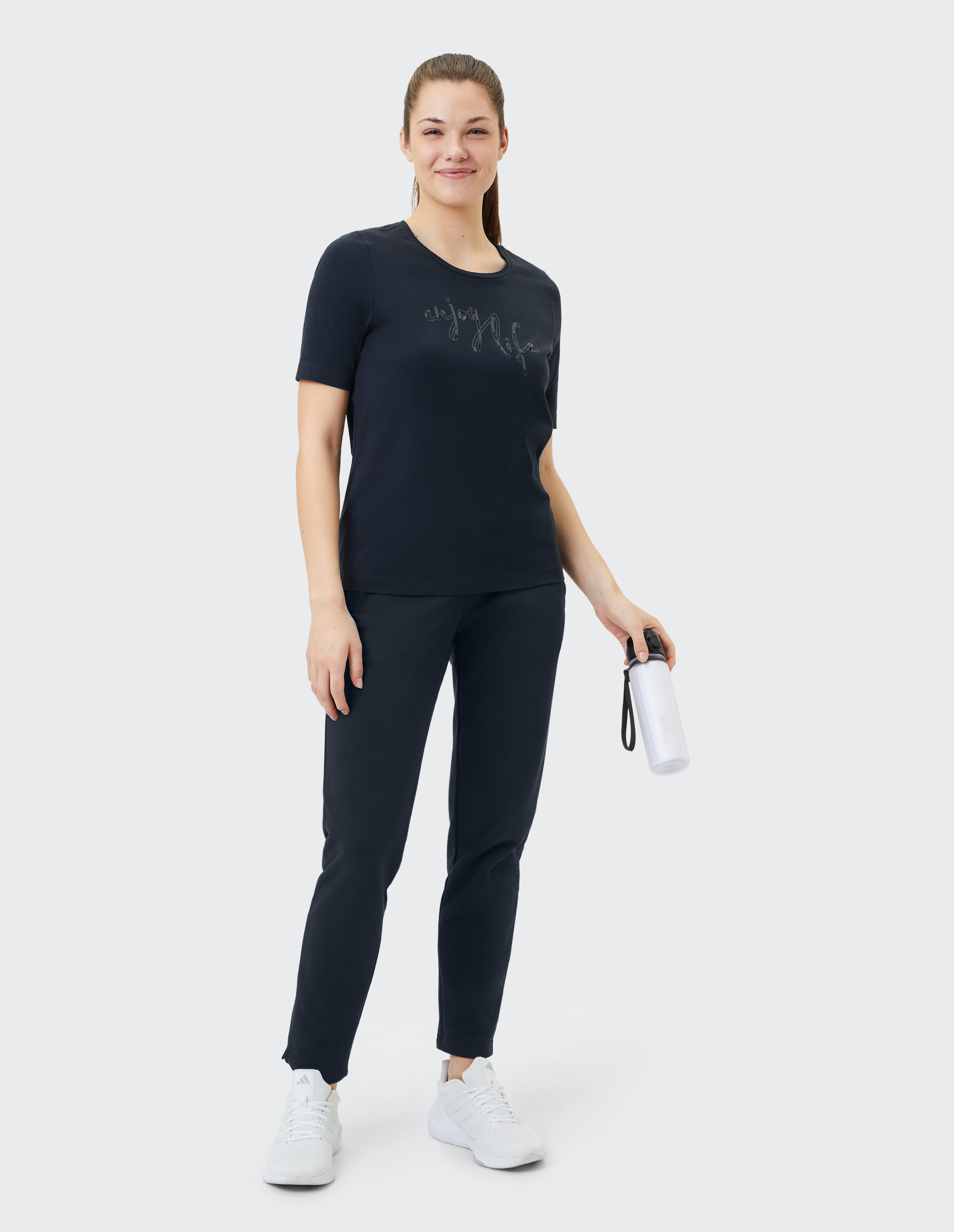 Frau in marineblauer Freizeitkleidung mit grafischem T-Shirt und Jogginghose, hält eine Wasserflasche in der Hand.