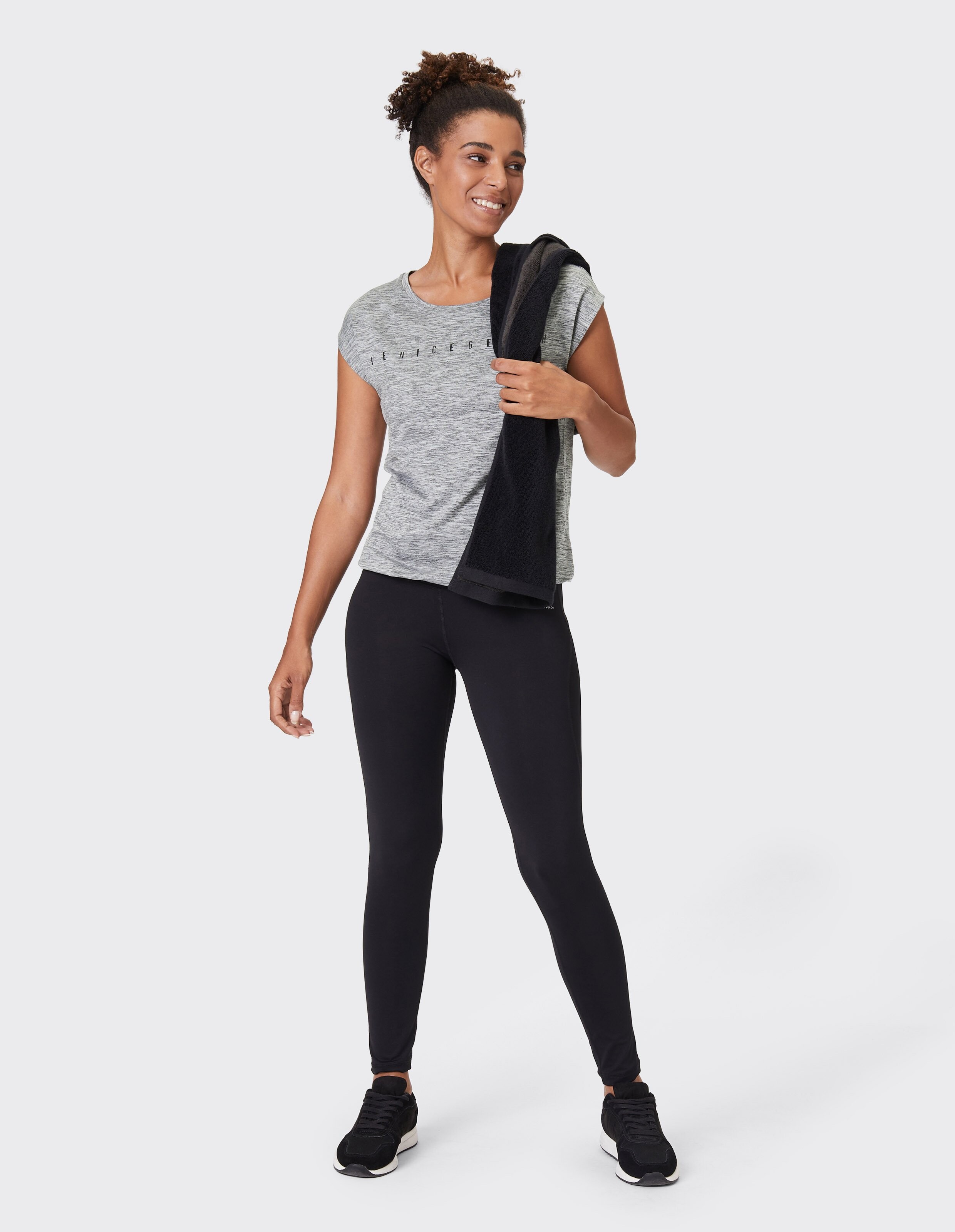 Frau in grauem T-Shirt und schwarzen Leggings hält ein Handtuch in der Hand und lächelt.