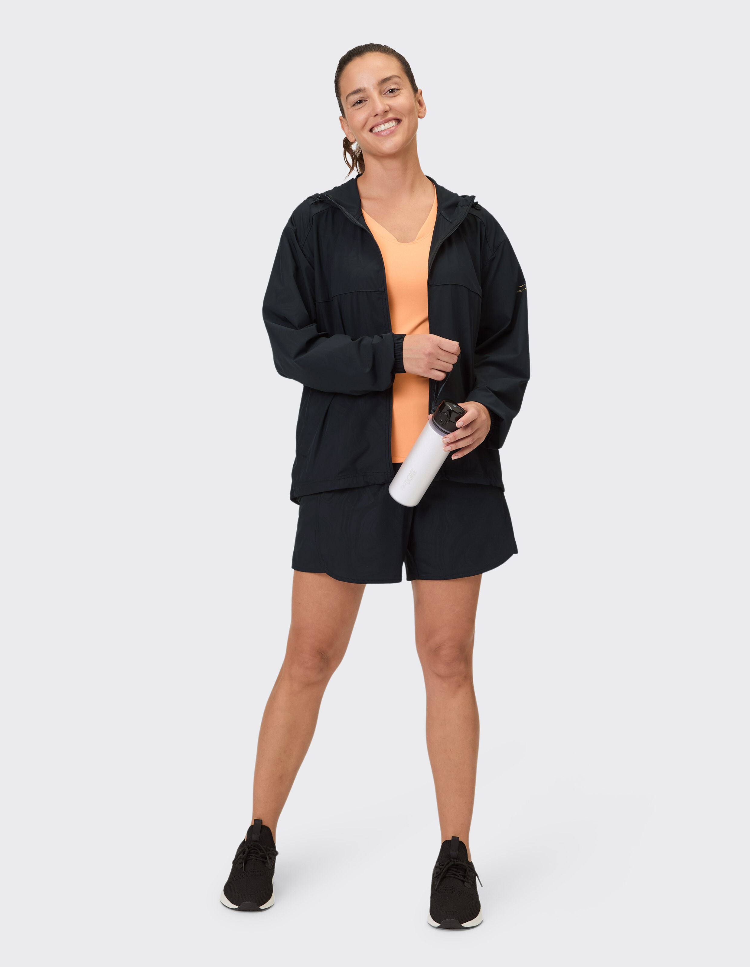 Frau in schwarzer Freizeitjacke und Shorts mit orangefarbenem Oberteil, hält eine Wasserflasche in der Hand.