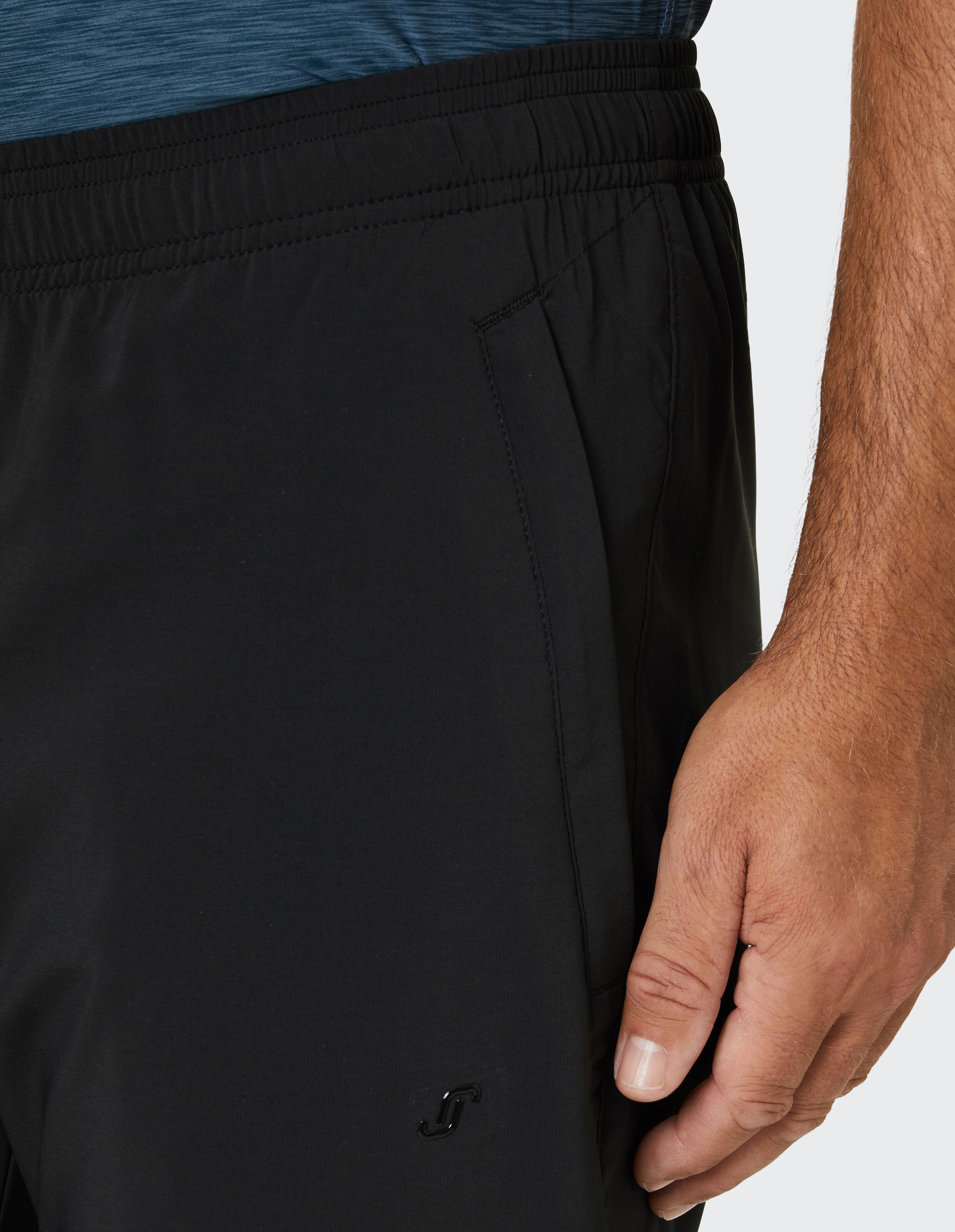 Nahaufnahme einer männlichen Hand, die neben einer schwarzen Hose mit elastischem Bund und dezentem Logo steht, vor blauem Shirt.