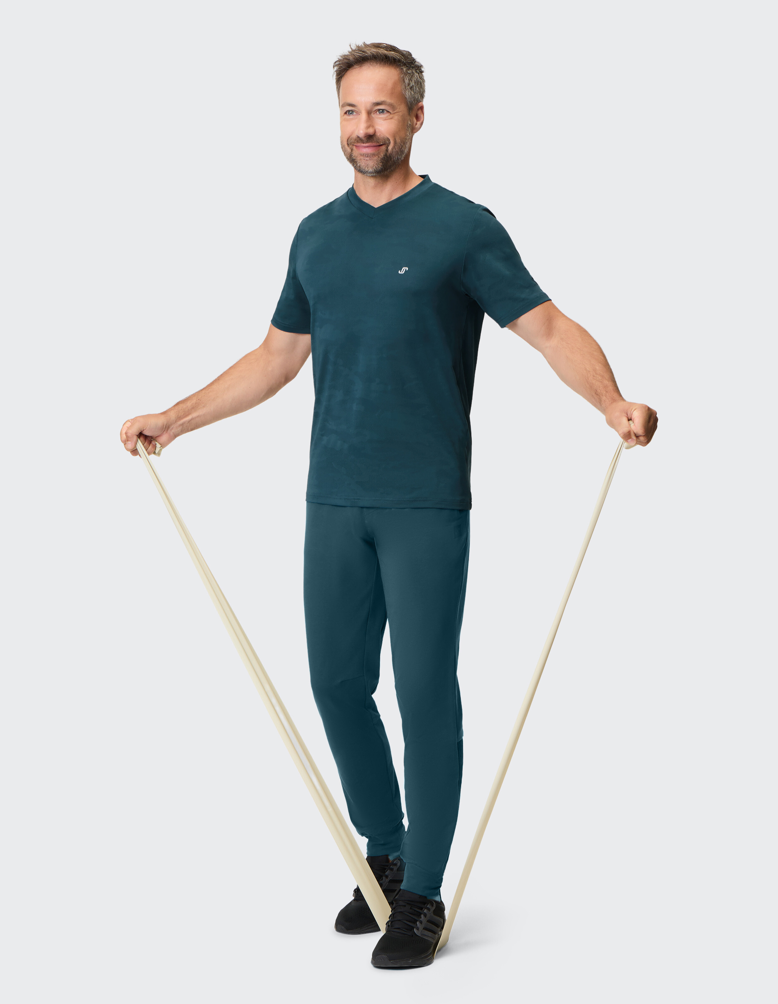 Mann in blaugrüner Freizeitkleidung mit T-Shirt mit V-Ausschnitt und taillierter Hose, der mit Widerstandsbändern trainiert.